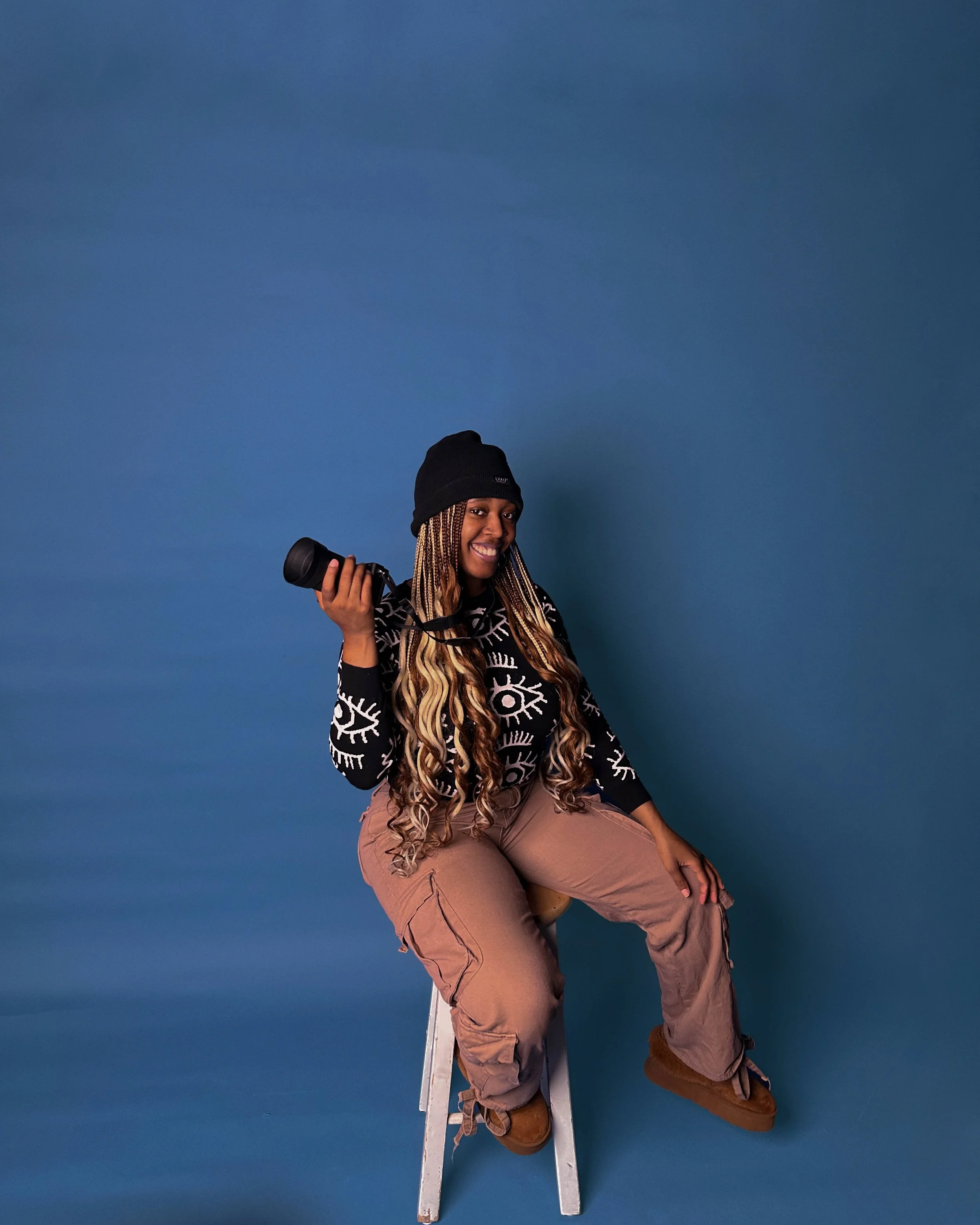 A woman with long, wavy hair sitting on a small ladder, holding a camera in her right hand, wearing a black beanie, a black sweater with white eye and eye socket designs, and tan cargo pants, smiling against a blue background.