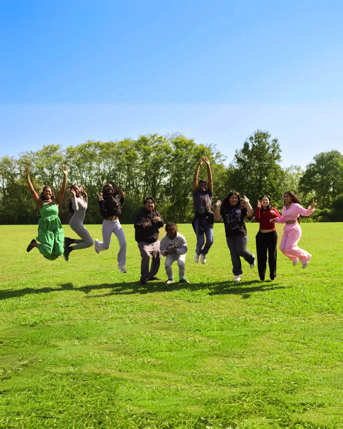 A group of nine diverse people are jumping and smiling on a grassy field with trees and a clear blue sky in the background.