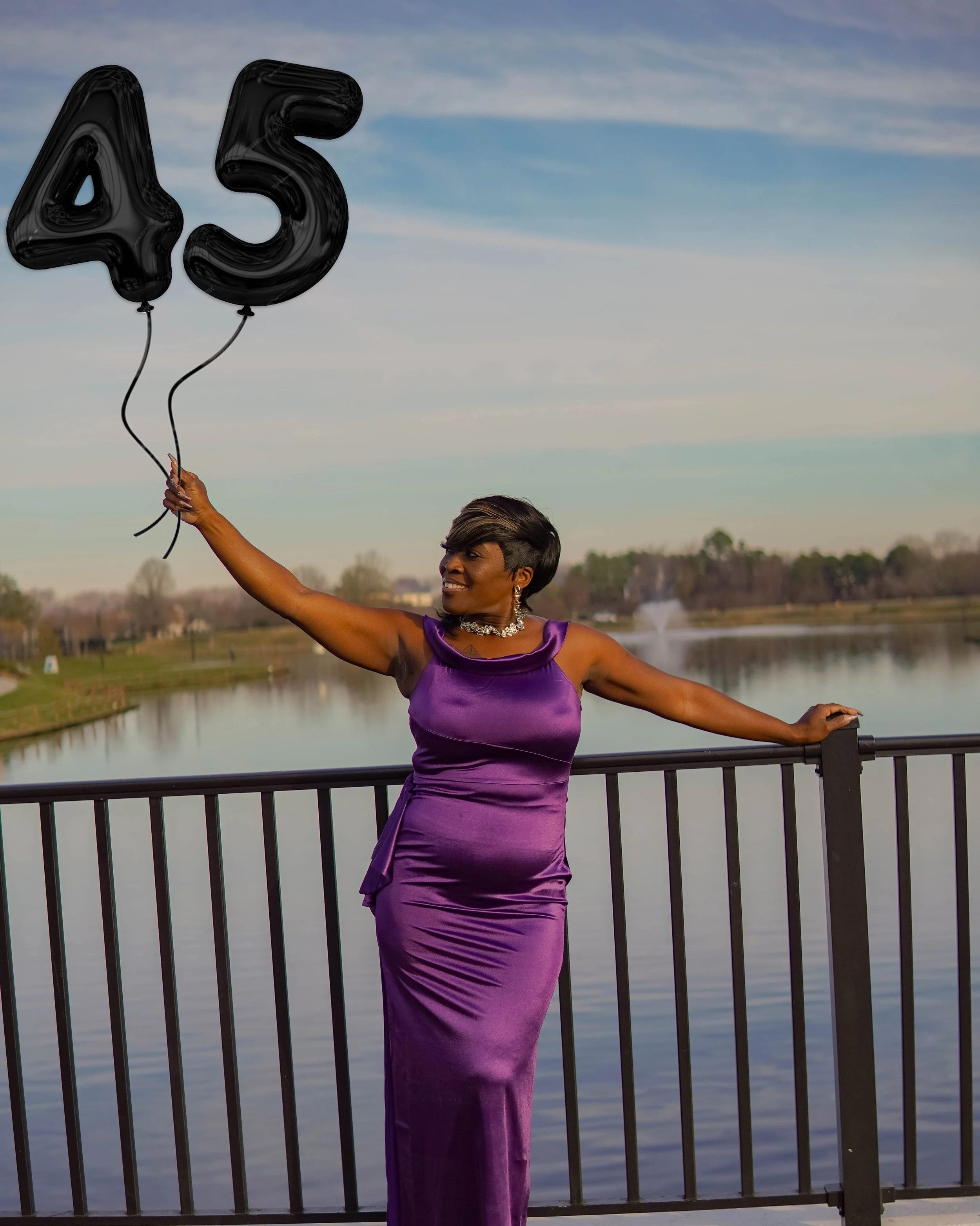 A woman in a purple satin dress holding two black balloons shaped as the numbers 4 and 5, standing on a bridge over a lake with a fountain, trees, and a cloudy sky in the background.