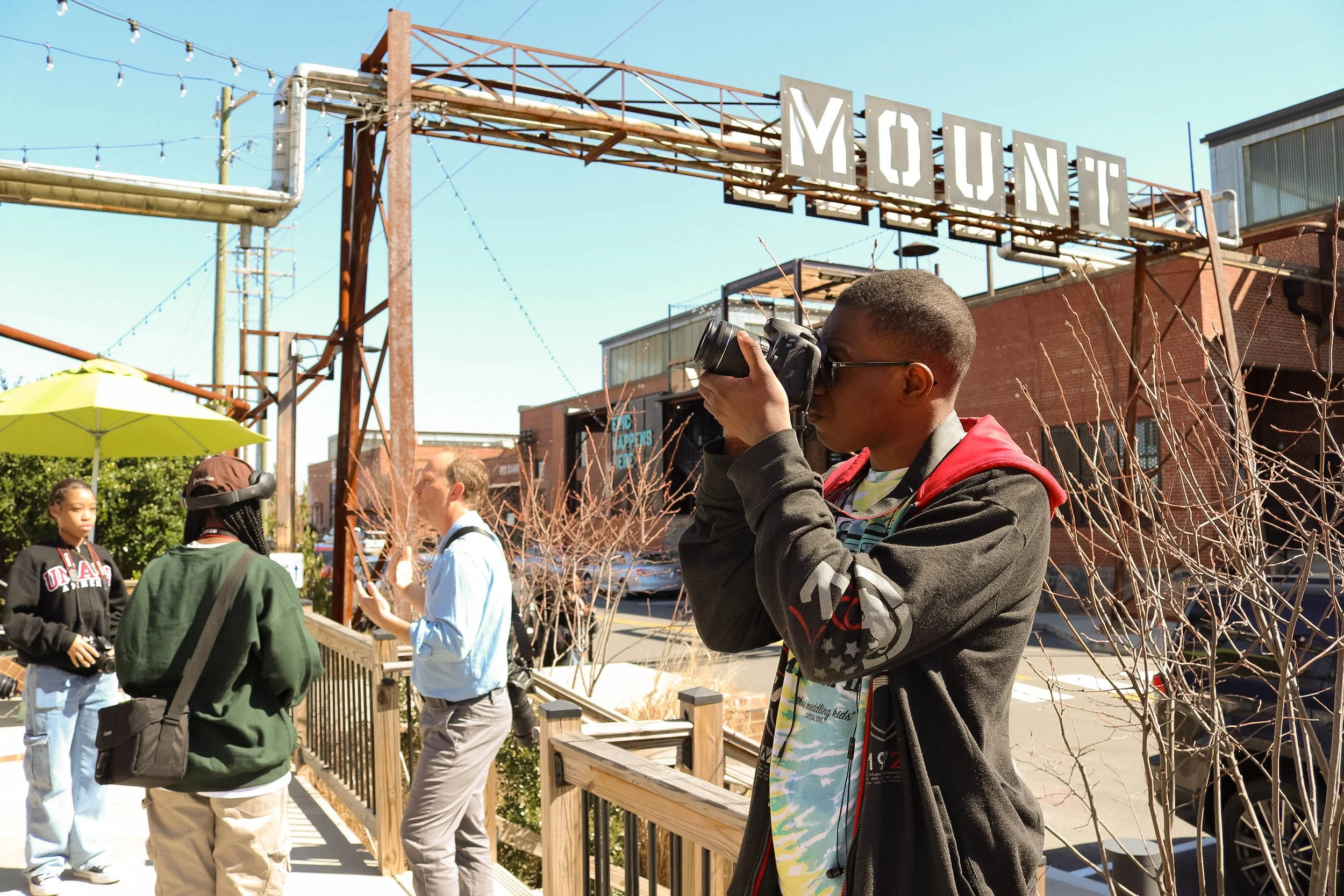 A young man with glasses and a dark jacket taking a photograph with a camera outdoors near a small crowd under a sign that reads 'MOUNT' on a clear sunny day.