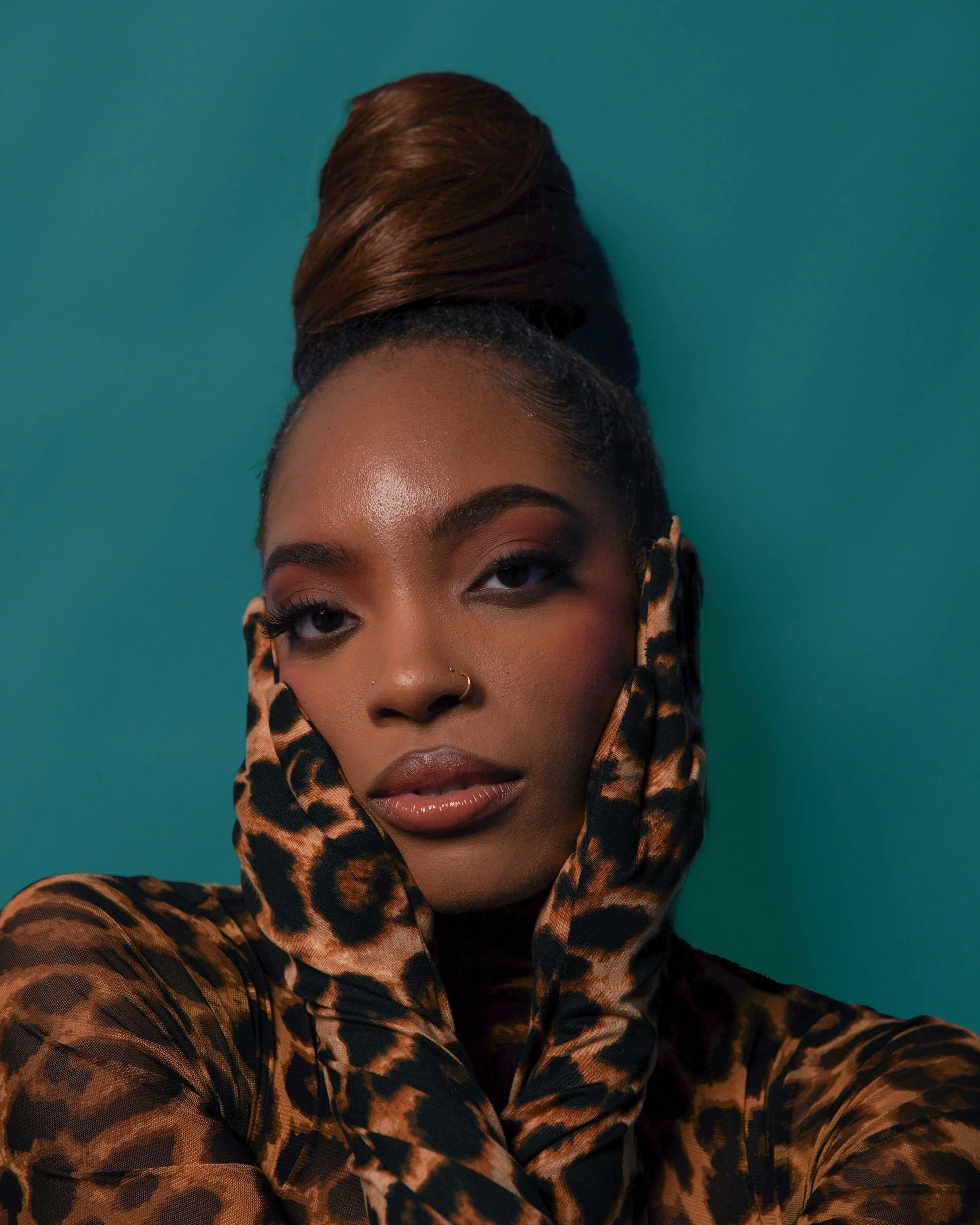 A woman with dark skin and styled hair, wearing leopard print gloves and clothing, posing against a teal background with hands on her face.