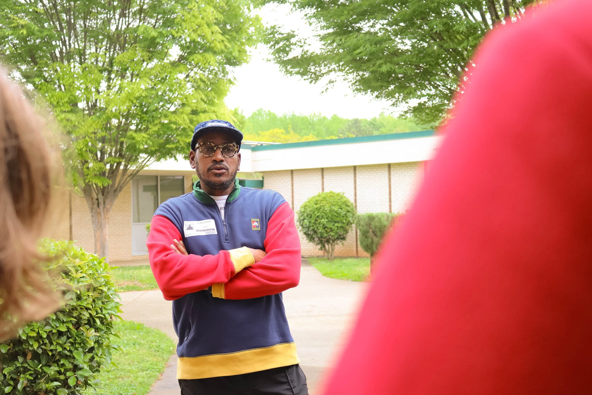 A man wearing glasses, a blue and red jacket, and a cap stands outdoors with arms crossed, during daytime, with trees and a building in the background.
