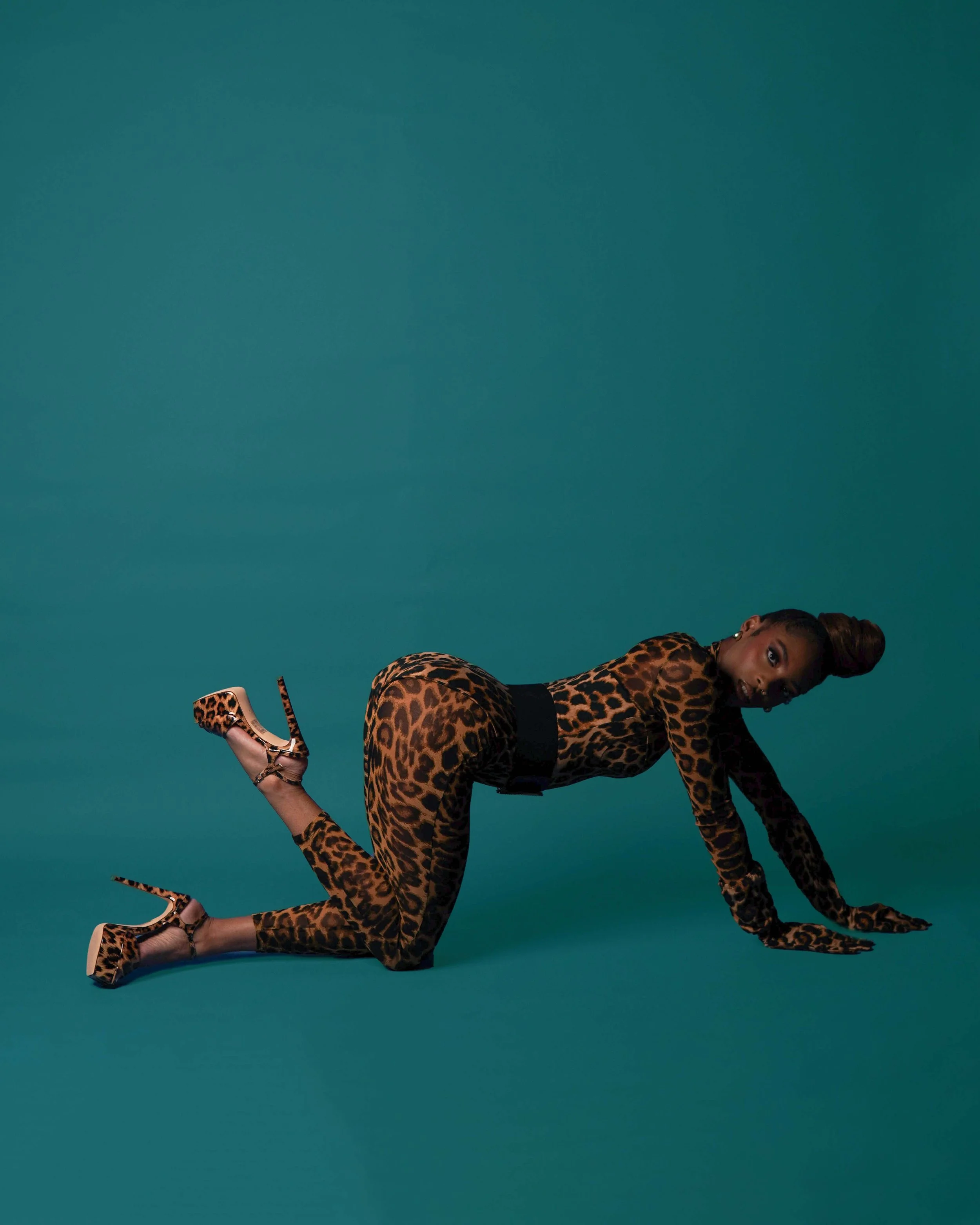 A woman on all fours wearing a leopard print outfit and matching heels, on a teal background.