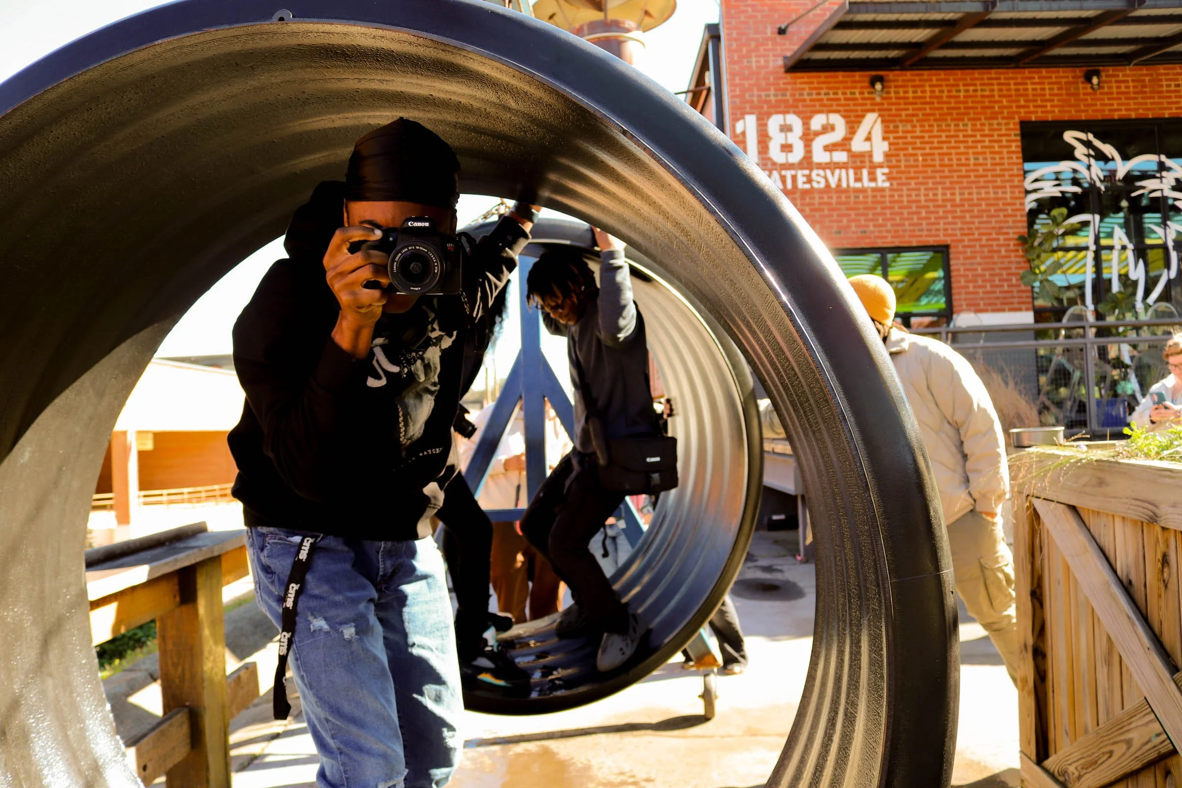 Person taking a photograph through a large, curved metal tunnel, with other people walking behind inside the tunnel. The background shows a brick building with the number 1824 and the word GATESVILLE.