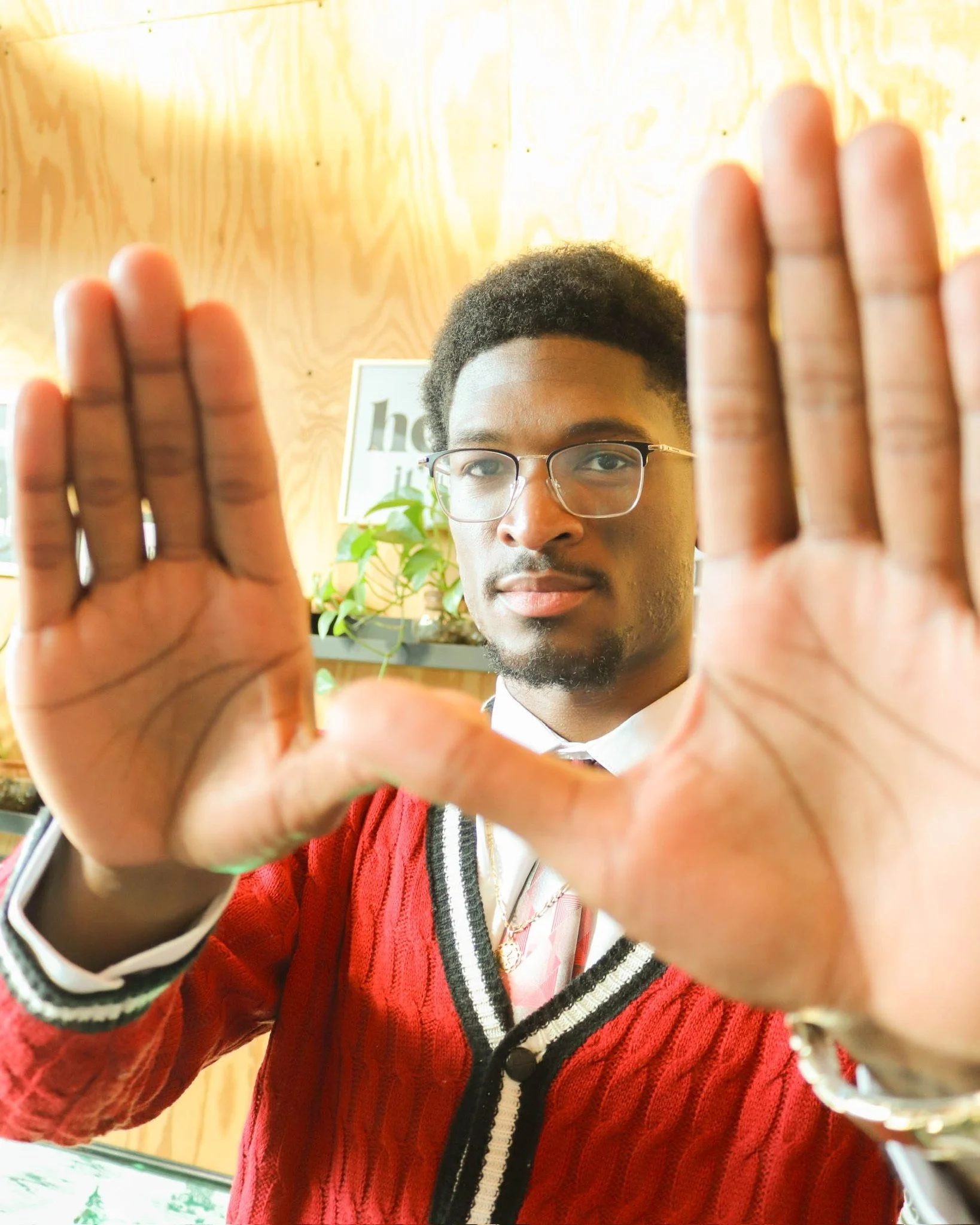 A young man with glasses and a red sweater with white and black accents holding his hands up to the camera, framing his face, in an indoor setting with wooden walls and some green plants.