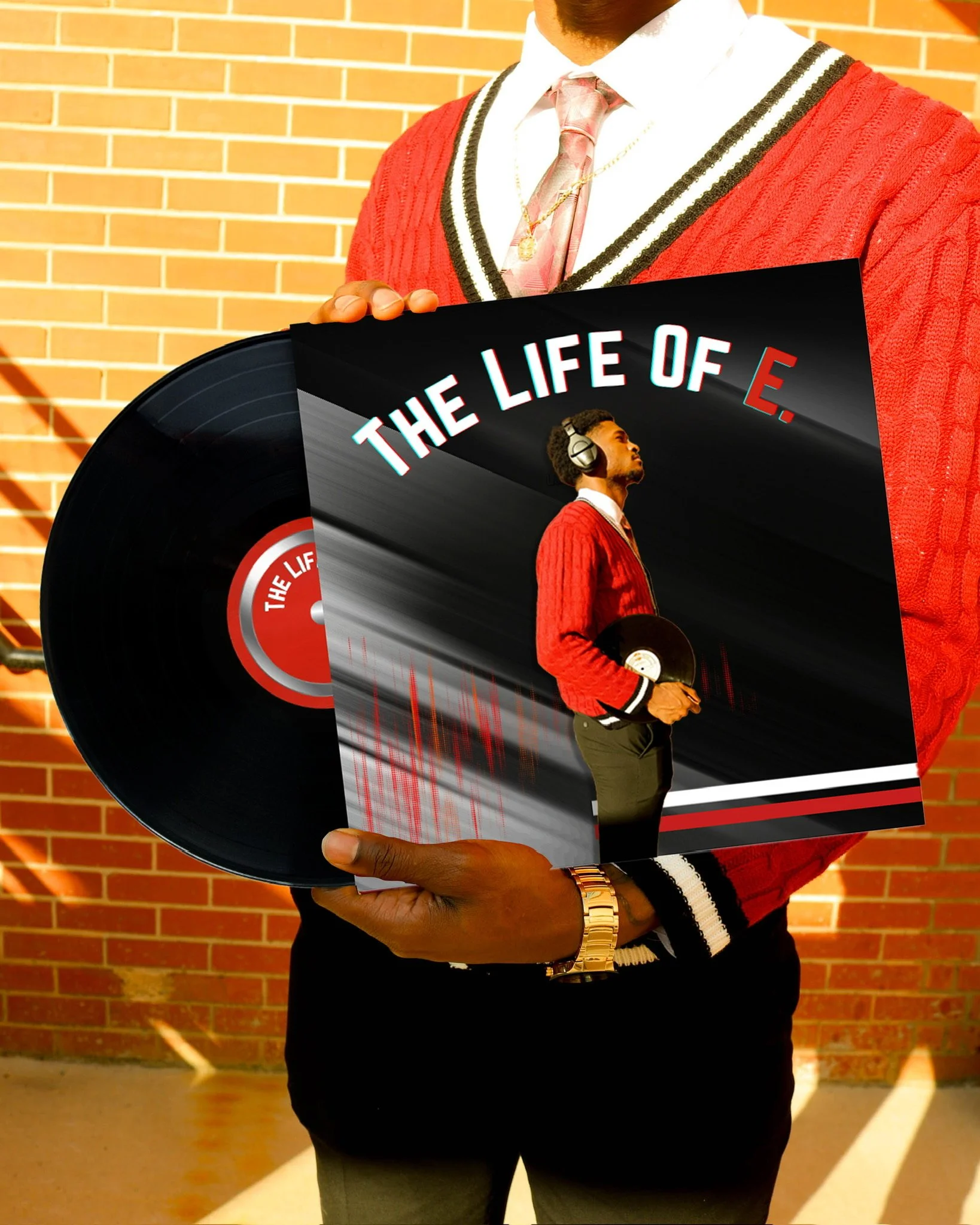 A man dressed in a red sweater, white shirt, and pink tie, holding a vinyl record and an album cover with the title 'The Life of E.' in front of a brick wall.