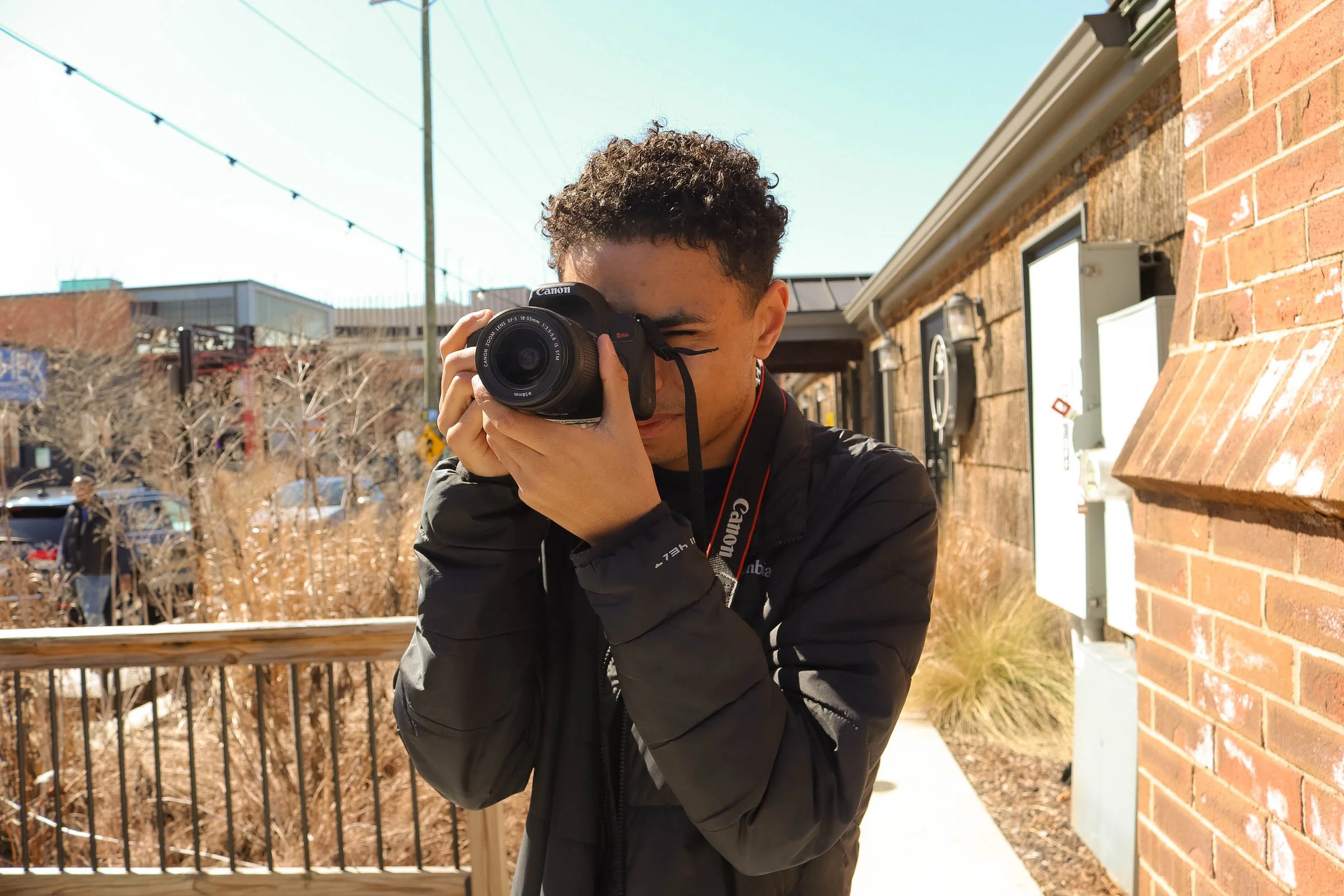 Young man with curly hair taking a photo with a Canon camera outdoors on a sunny day, standing next to a brick building.