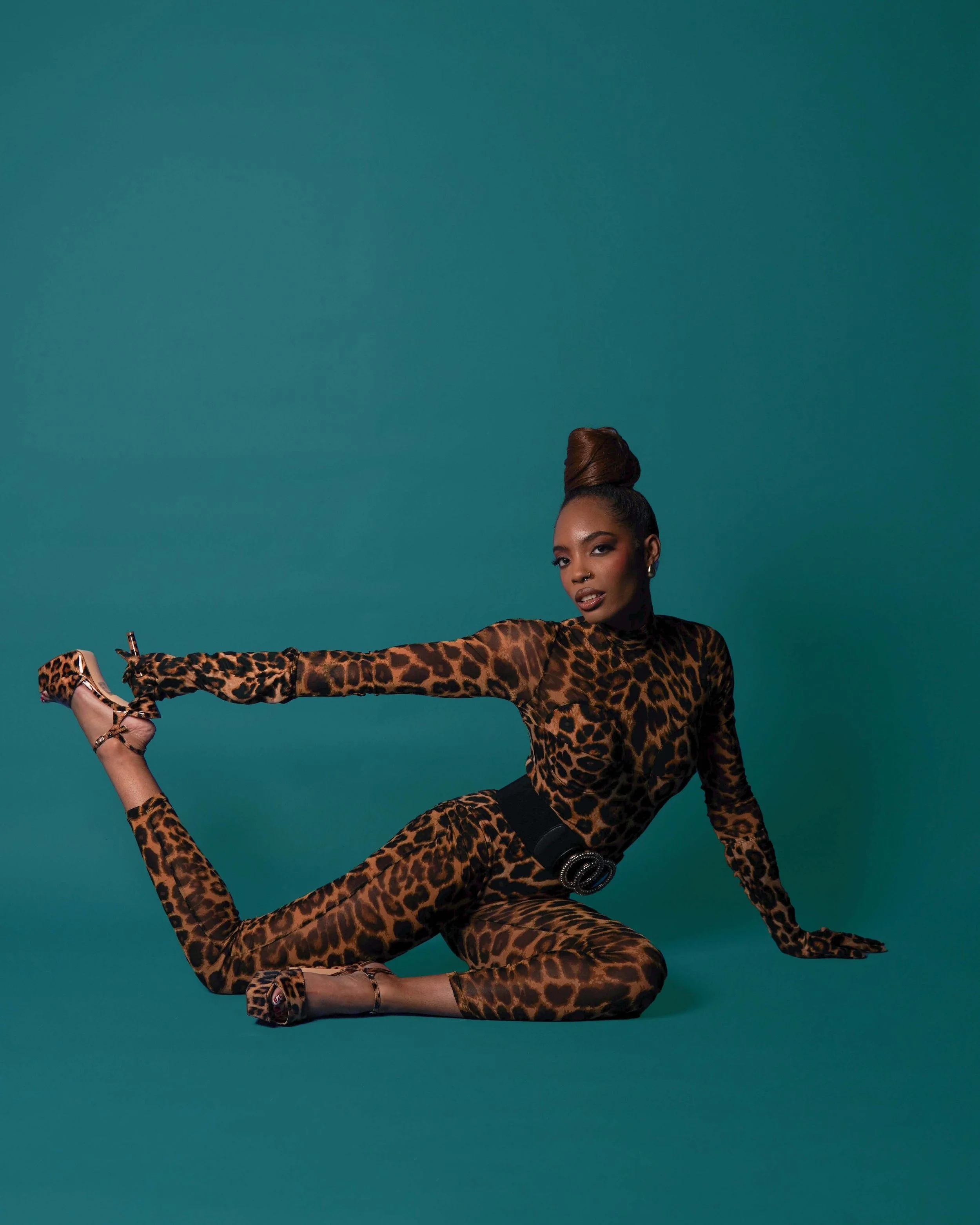 A woman wearing a leopard print jumpsuit, high heels, and a black belt, posing against a teal background with one hand on the ground and one leg extended.