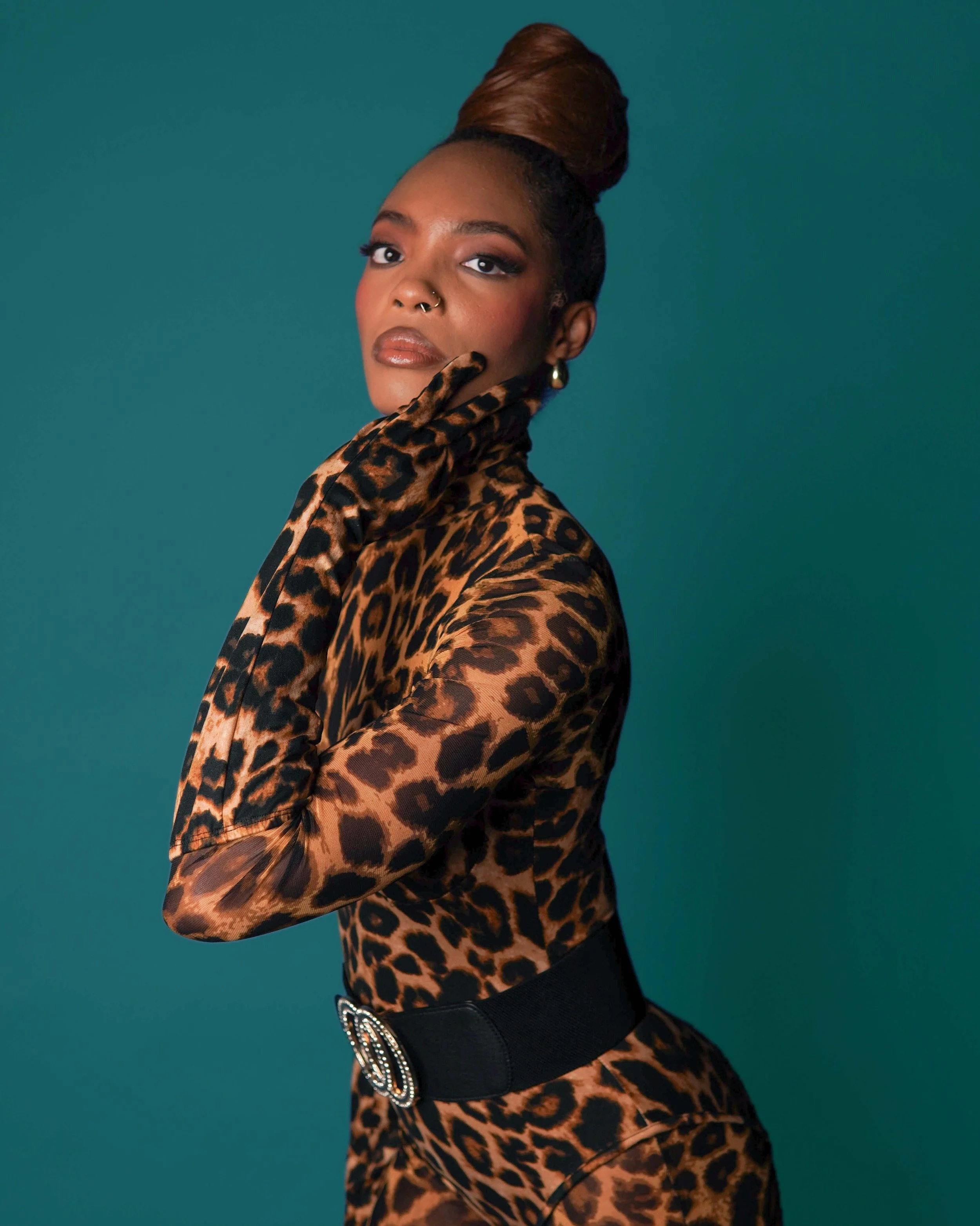 A woman in a leopard print outfit with matching gloves and a black belt with a large decorative buckle, posing against a teal background.