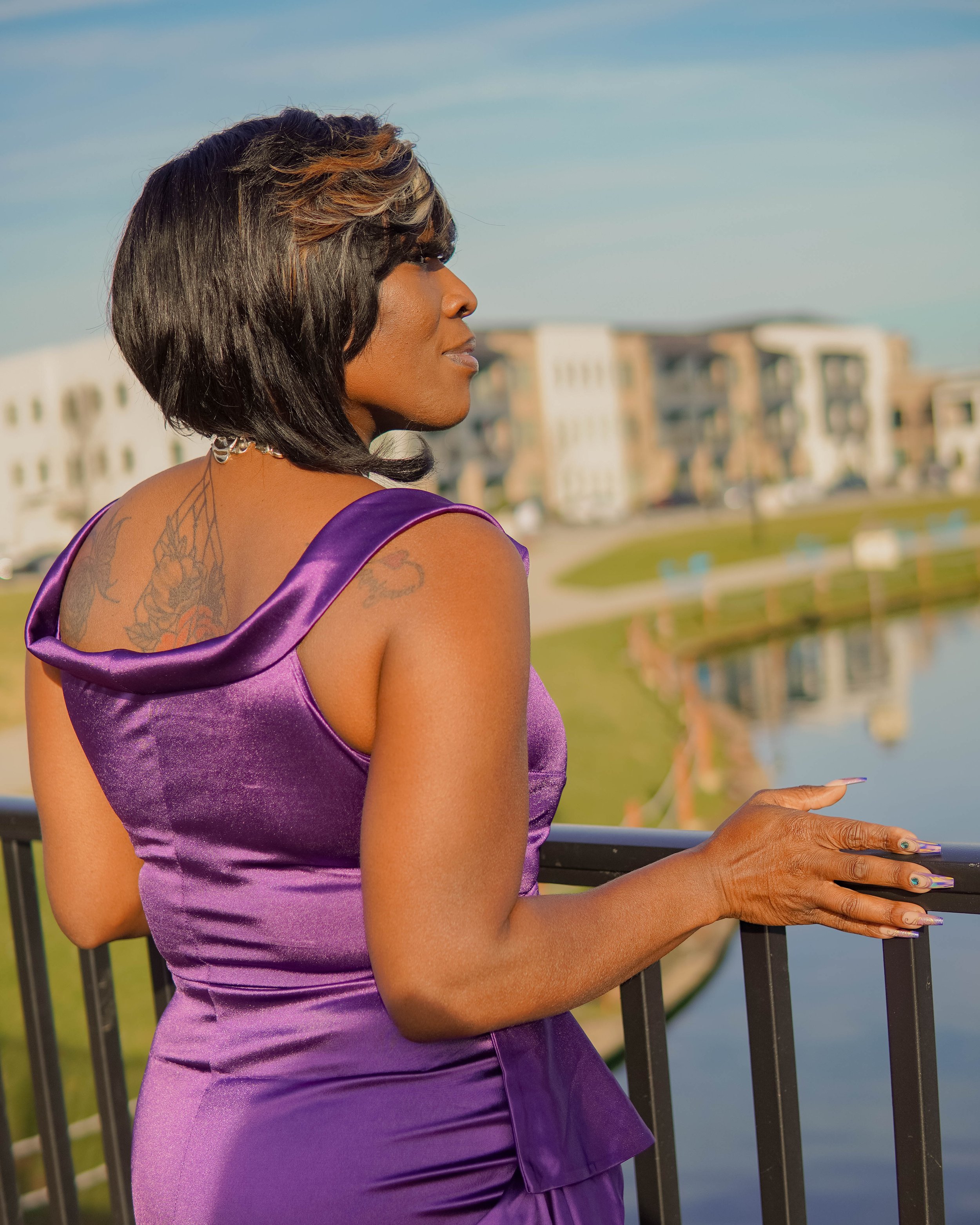 A woman with short black hair and a haircut with brown highlights, wearing a purple satin dress, standing by a black metal railing near a body of water with buildings in the background.