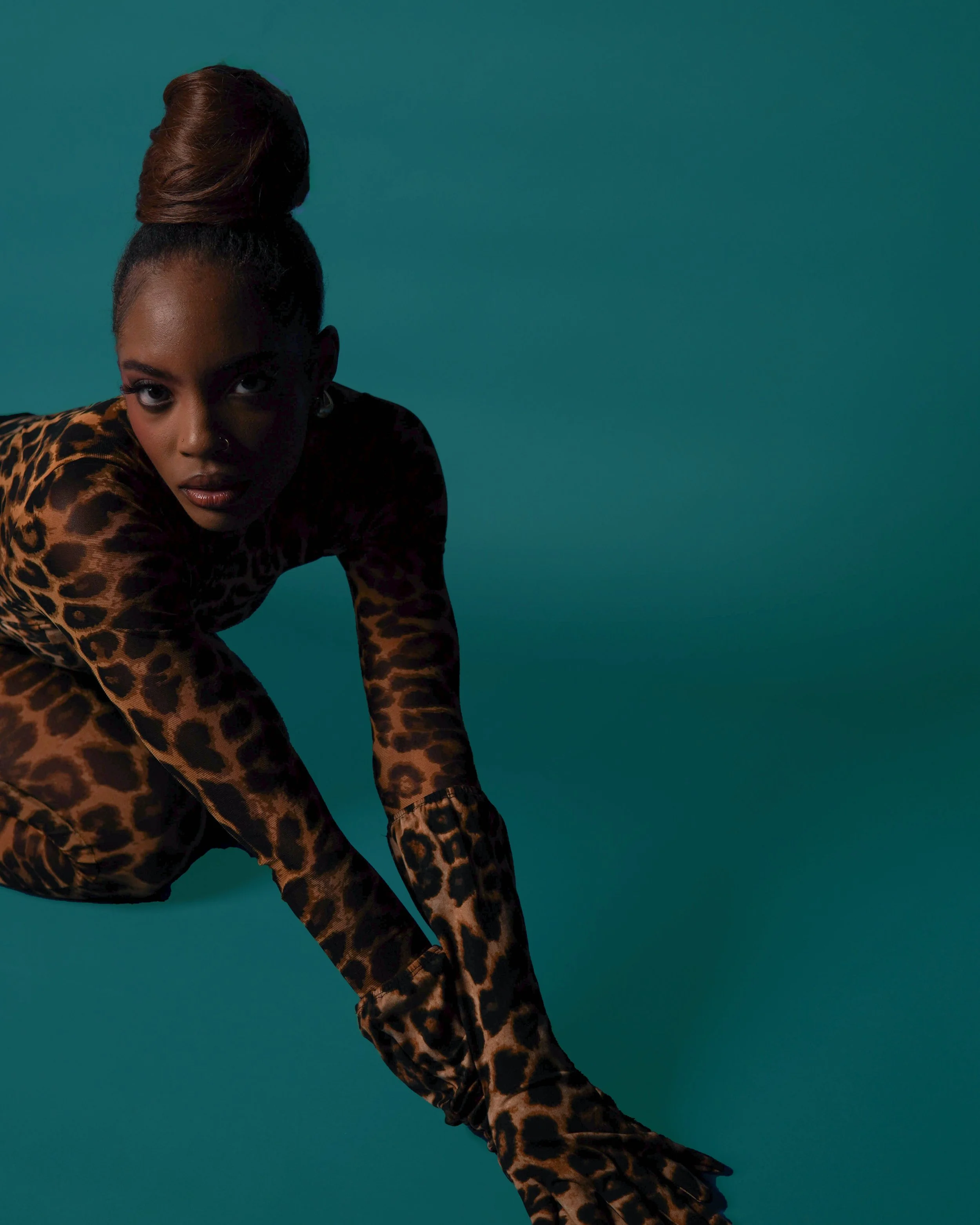 A woman in leopard print clothing crouching against a solid teal background, facing the camera with intense expression.