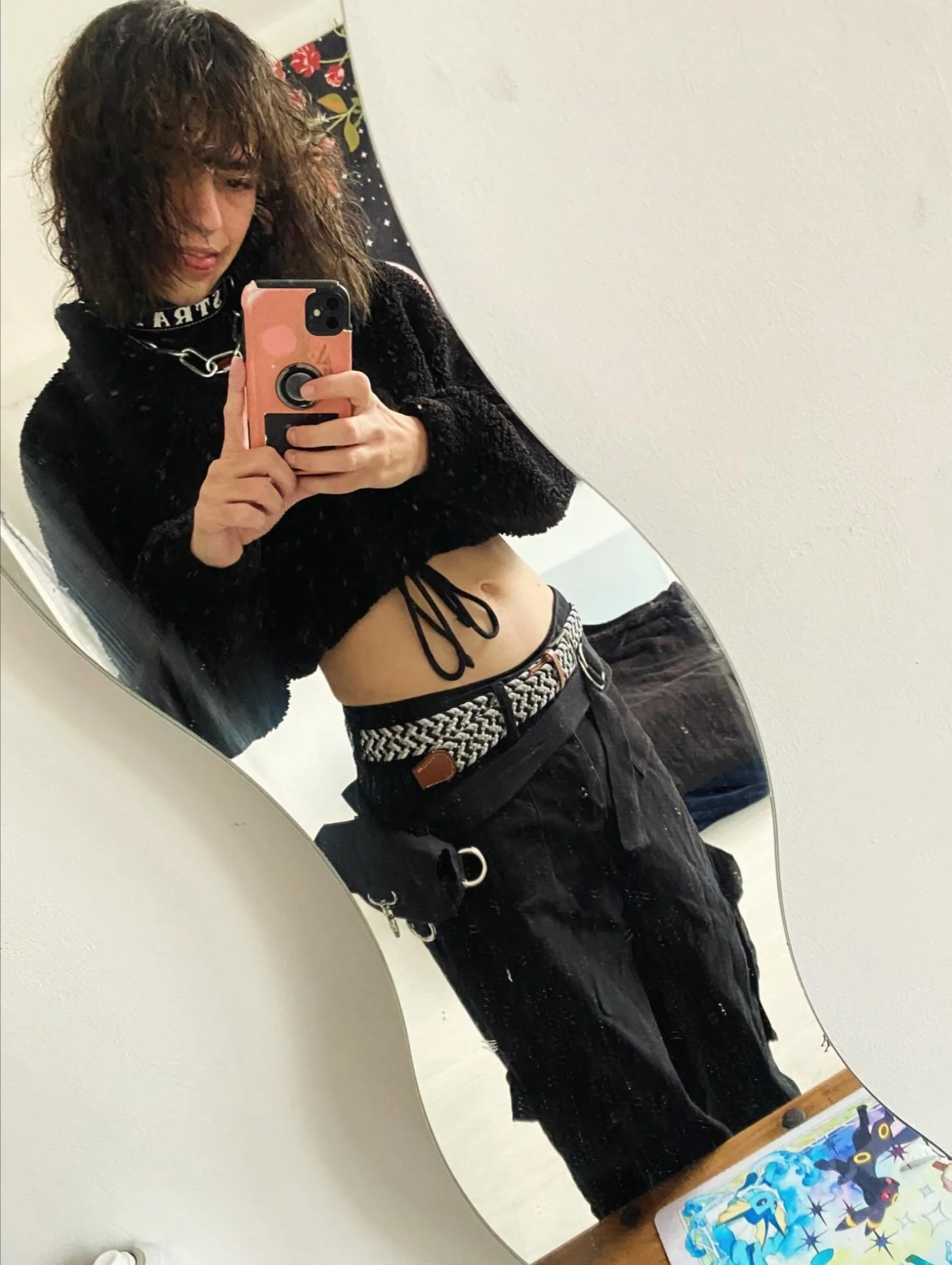 A person taking a mirror selfie in a room with a wavy mirror, showing their midriff, wearing a black cropped hoodie, black pants, a woven belt, and holding a pink phone.