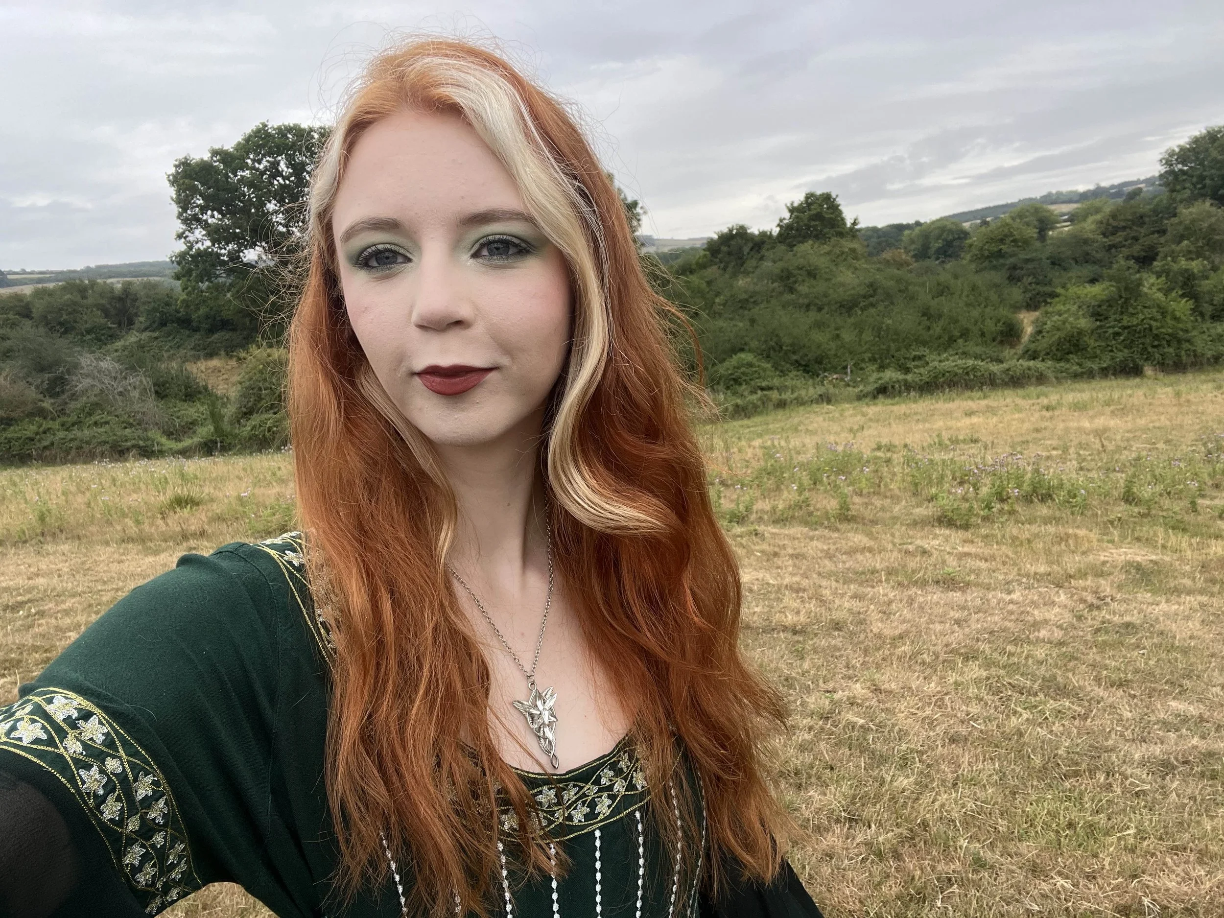 A woman with long red hair and makeup taking a selfie outdoors in a grassy field with trees and hills in the background.