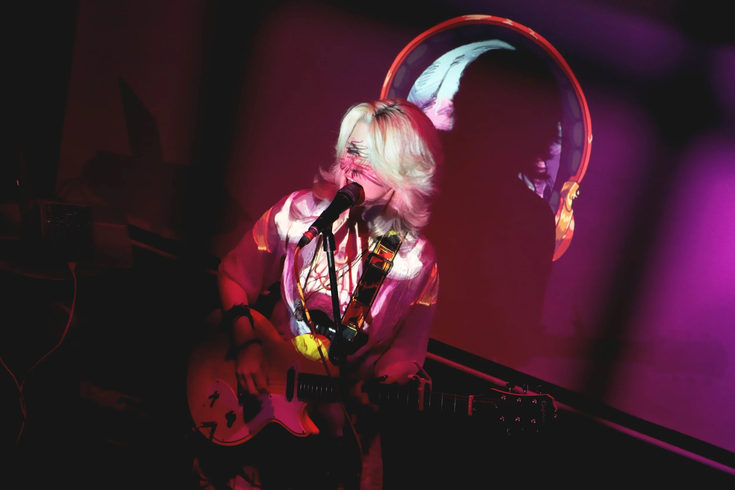 A person with blond hair playing an electric guitar and singing into a microphone during a live performance, with colorful lighting and a large circular speaker in the background.