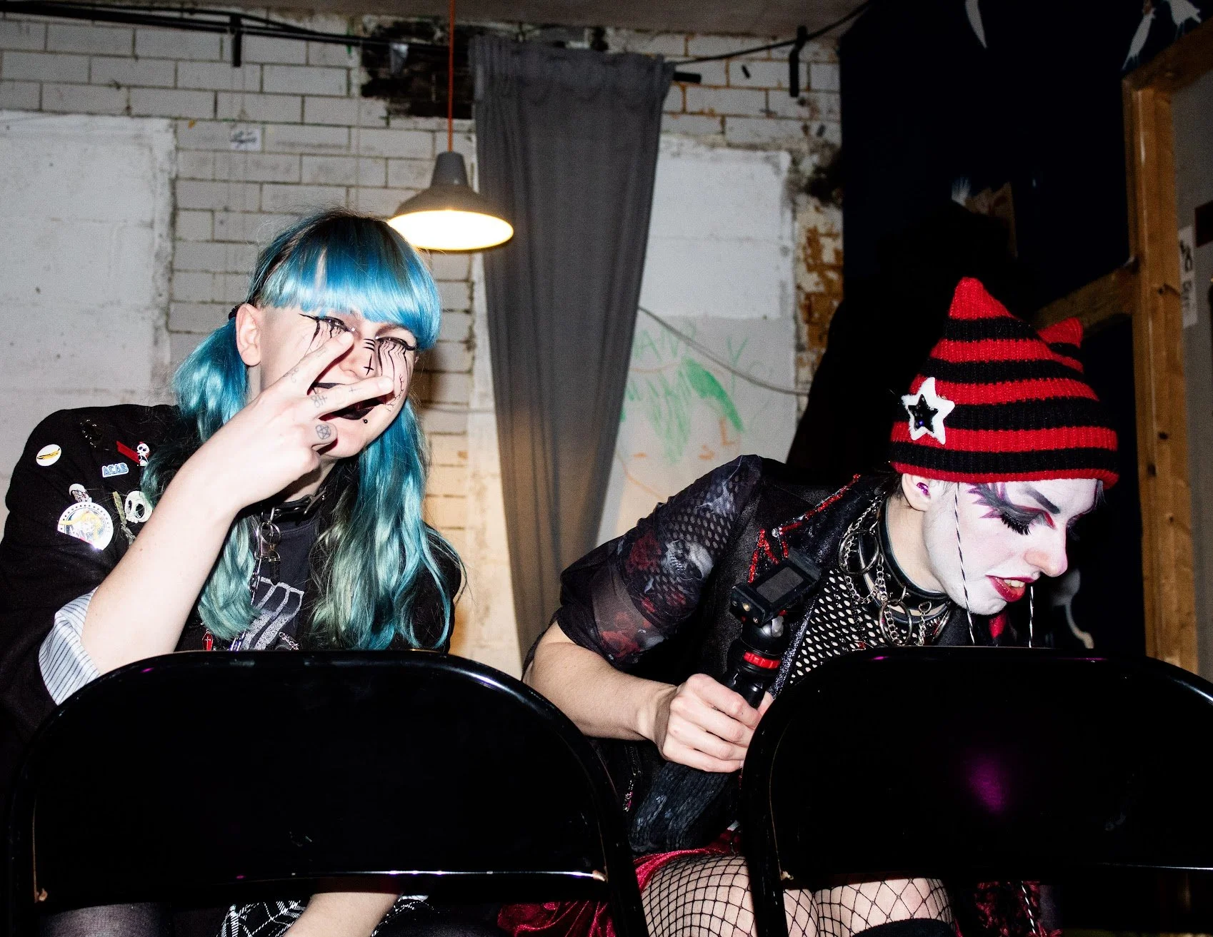 Two women with alternative fashion seated next to each other, one with blue hair making a peace sign and the other with a red and black striped beanie holding a microphone, both dressed in punk-inspired clothing, in a dimly lit indoor setting.