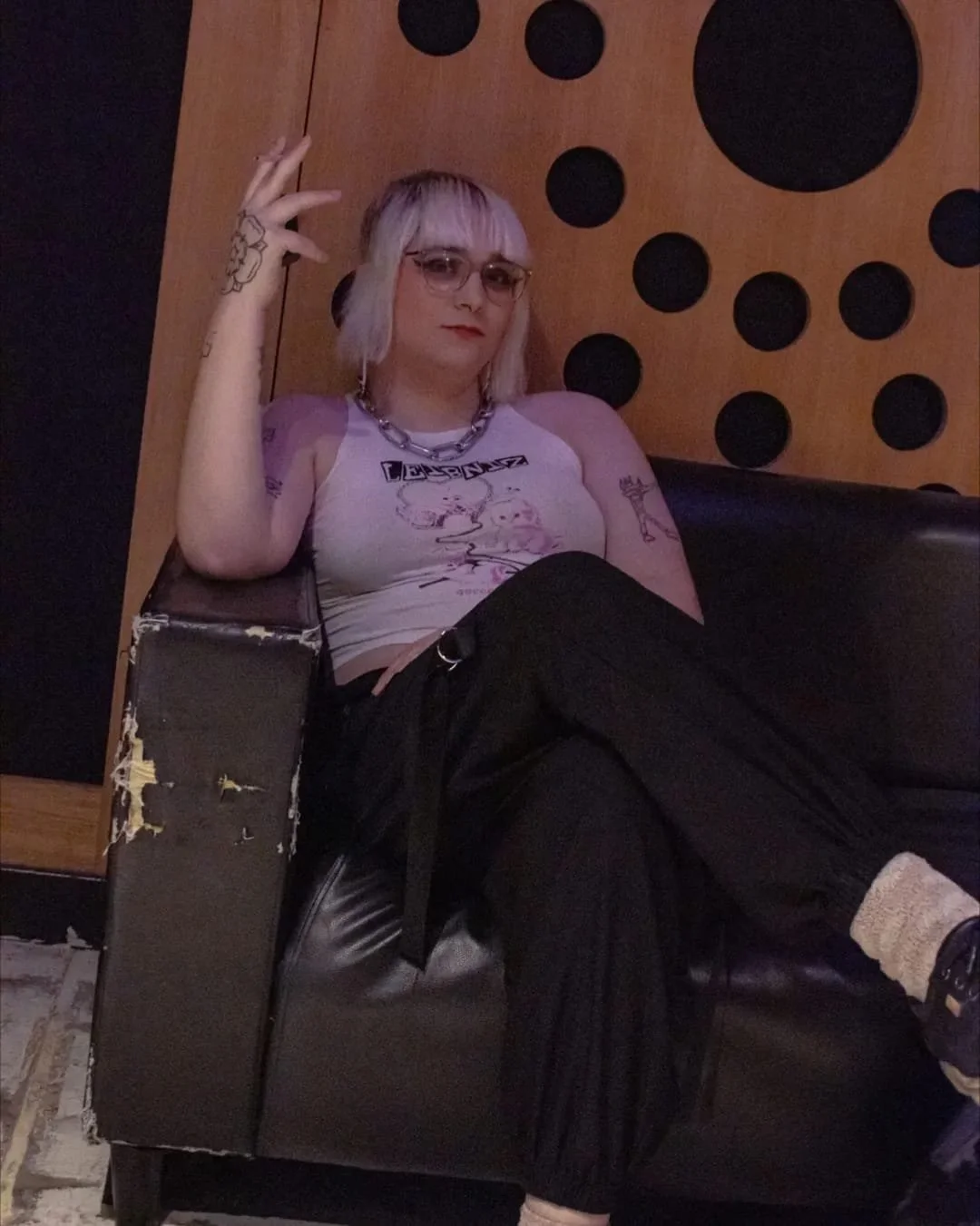 A young woman with pastel pink and purple hair, wearing glasses, a white graphic tank top, black pants, and patterned socks, sitting on a leather couch in a room with a wooden wall decorated with black circles, posing with her right hand raised and fingers in a peace sign.