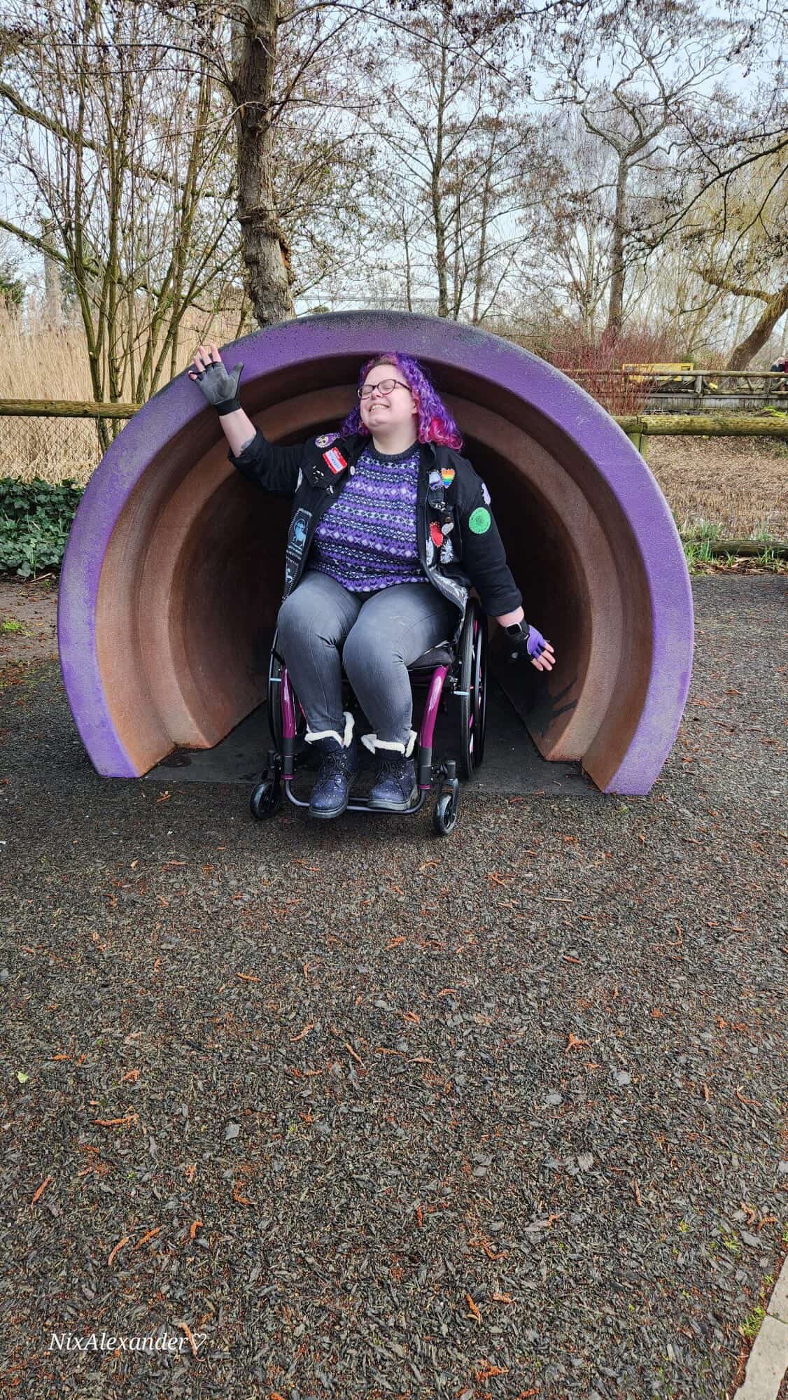 Person with purple hair and glasses sitting in a wheelchair at the entrance of a large purple and brown tunnel-shaped playground structure outdoors, smiling and waving.