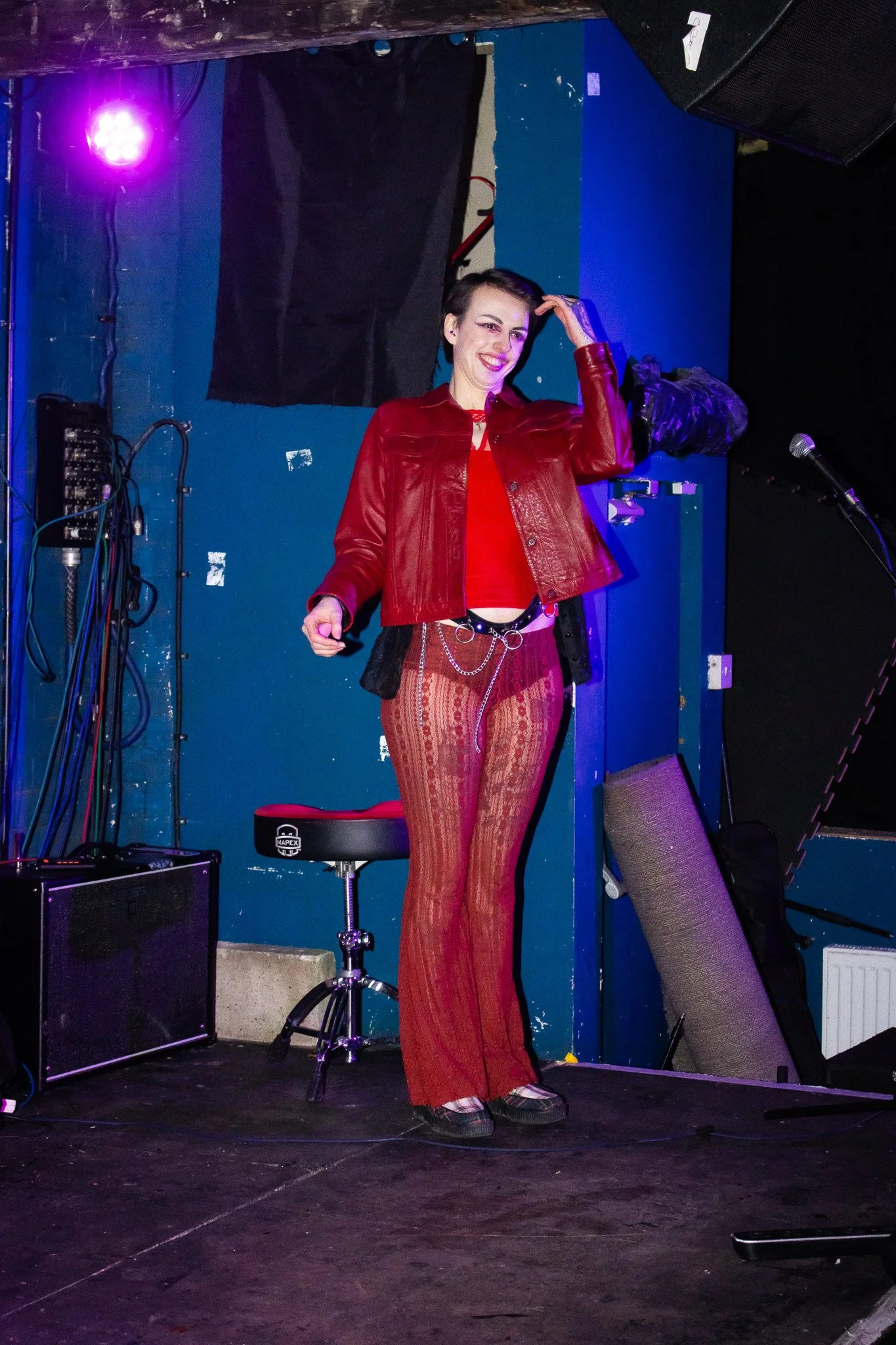 A woman on stage in a red leather jacket, red top, and patterned red pants, smiling and touching her hair, with music equipment and a microphone nearby.