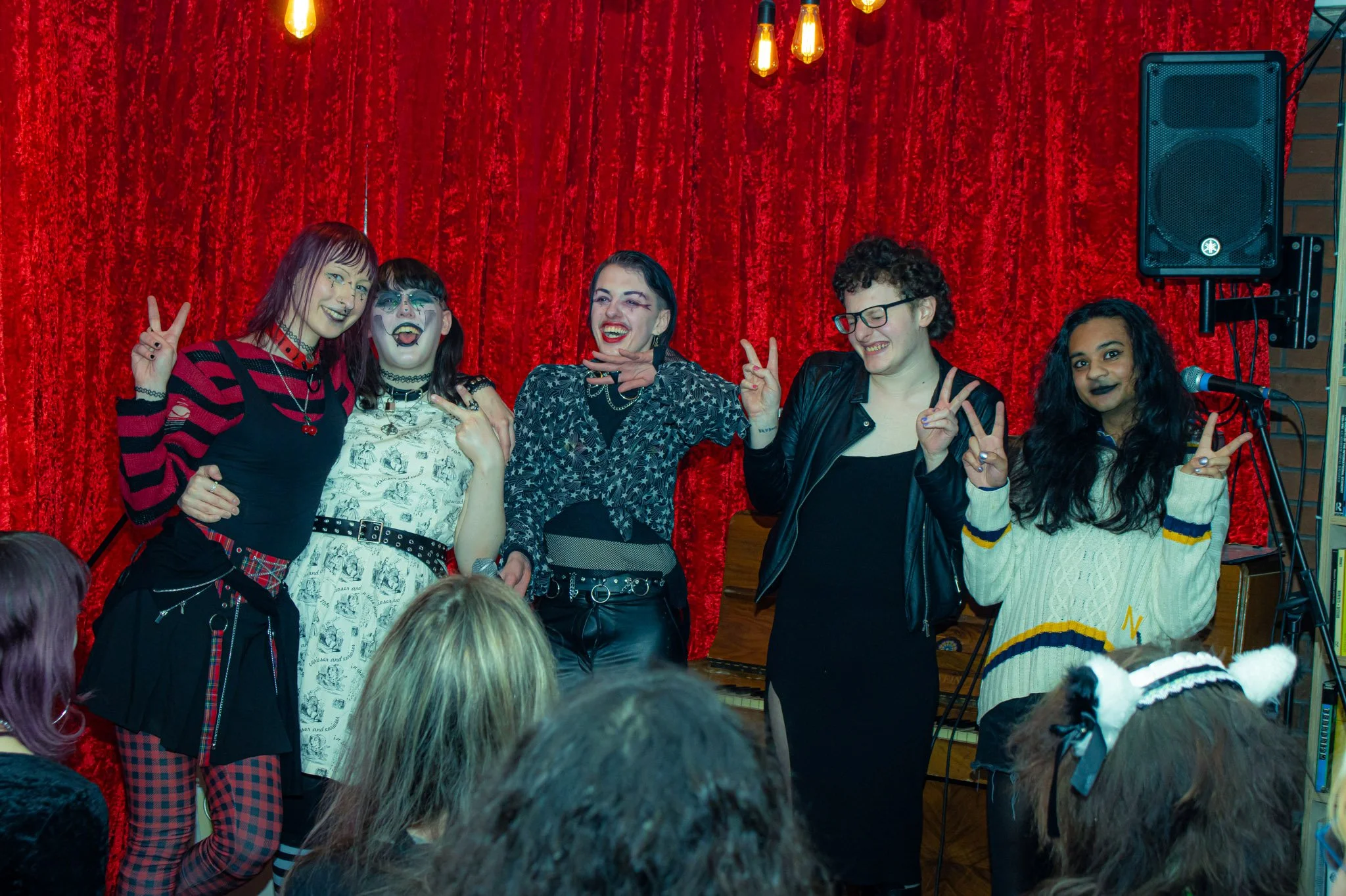 Group of six women performing on stage in front of a red curtain, making peace signs and smiling, with audience members in the foreground.