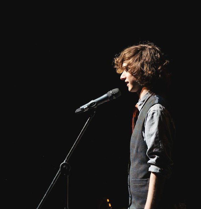 Edd, a non-binary individual, with curly hair standing in profile at a microphone on a stand, set against a black background.