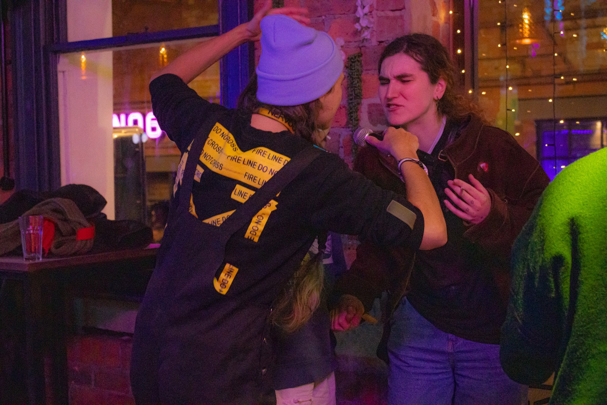 Two women are singing into a microphone in a dimly lit bar or club. One woman wears a gray beanie and a black jacket with yellow tape reading 'FIRE LINE DO NOT CROSS,' and the other woman has curly hair and wears a black jacket. They are close to each other, singing passionately.