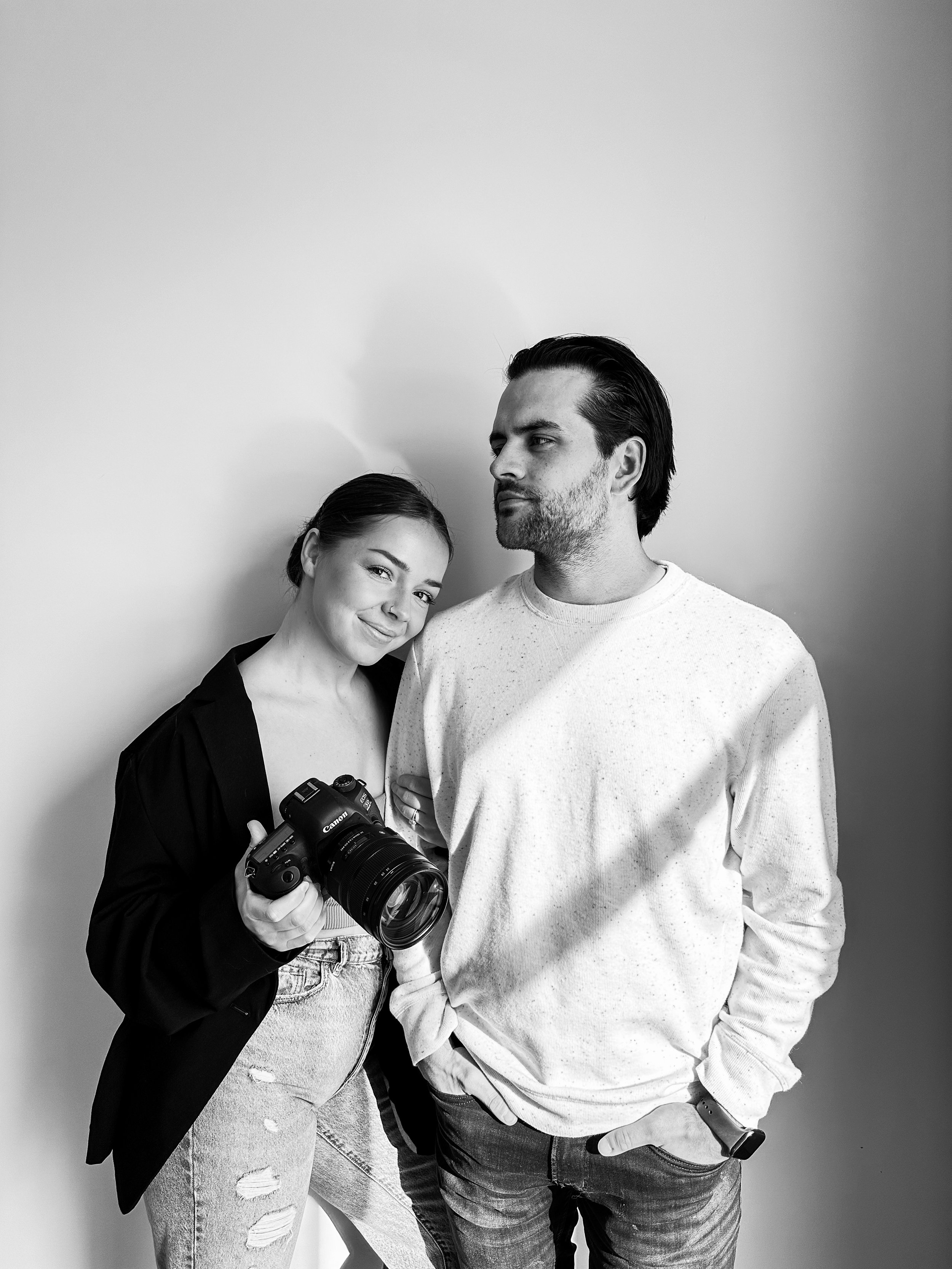 Black and white photo of a woman holding a camera, standing next to a man who has his hand in his pocket, both standing against a plain wall.