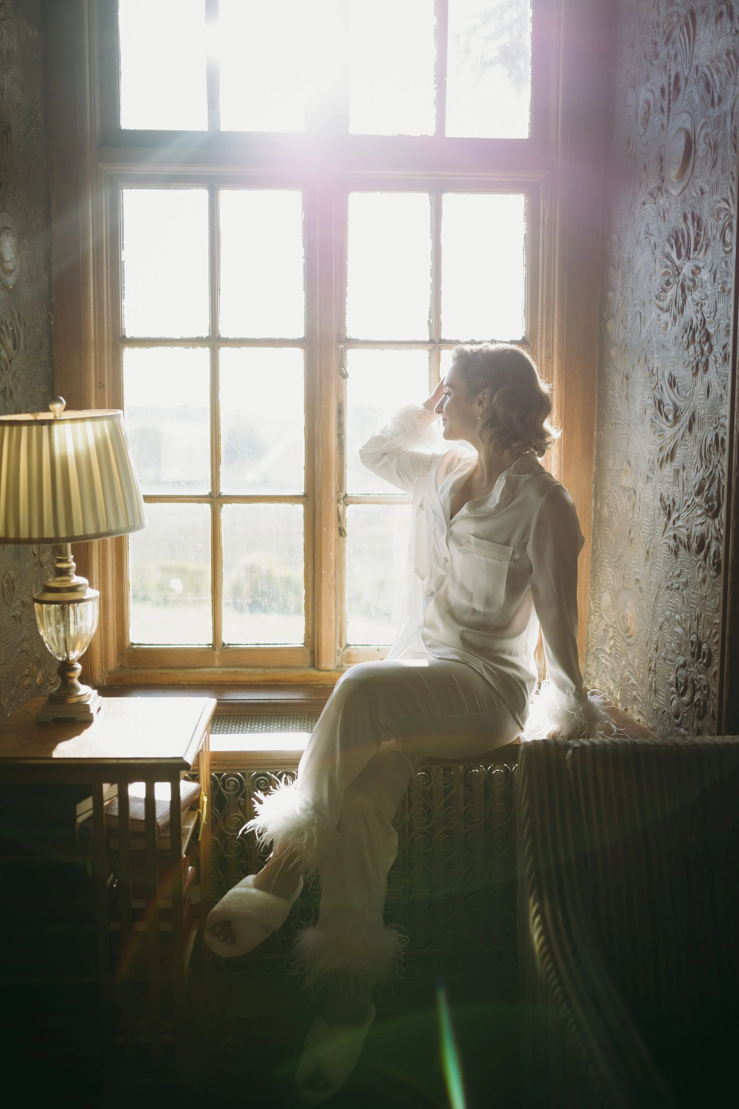 A woman sitting on a window seat in pajamas, backlit by sunlight coming through a large window, with a lamp on a small table beside her.