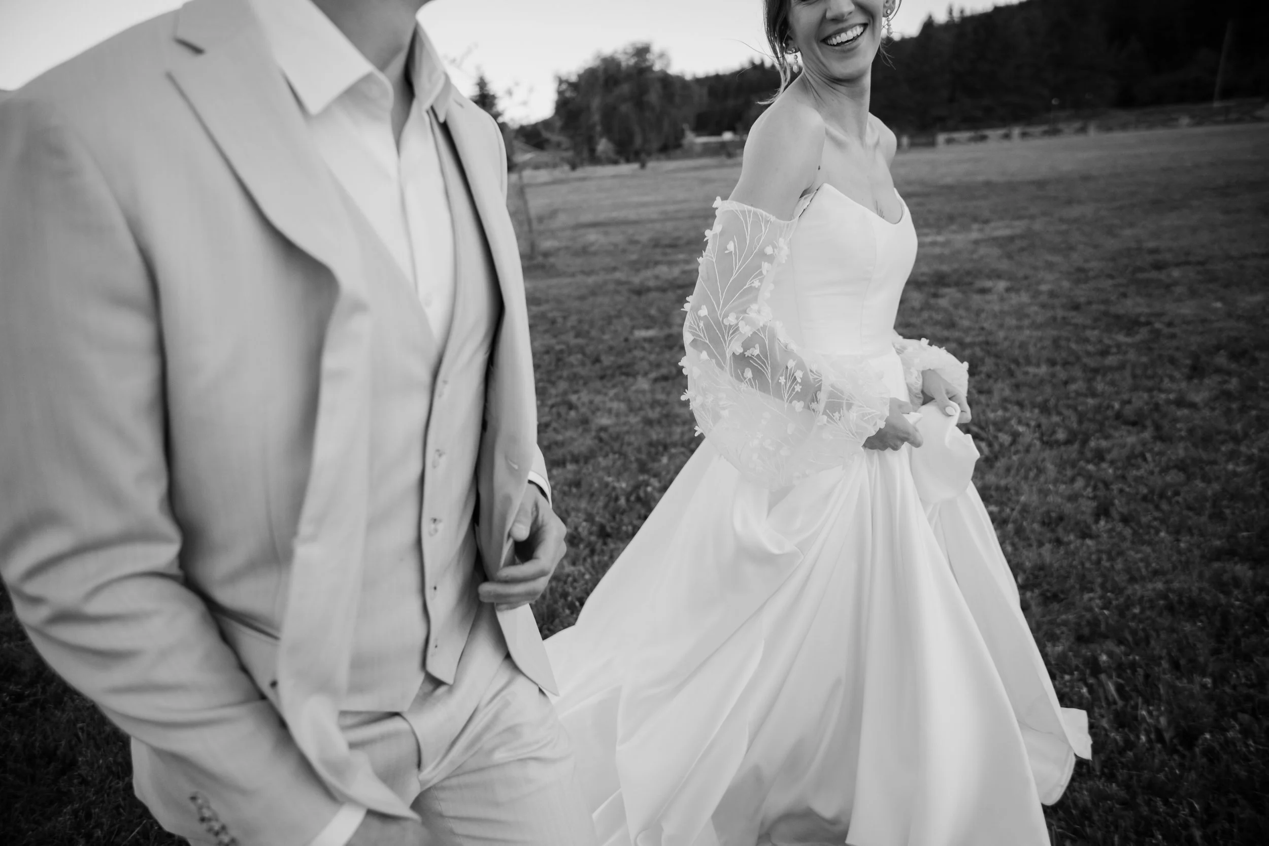 A black and white photo of a bride and groom walking outdoors on a grassy field. The groom is wearing a light-colored suit with his hands in his pockets. The bride is smiling, wearing a strapless wedding dress with lace off-the-shoulder sleeves, and holding part of her dress.