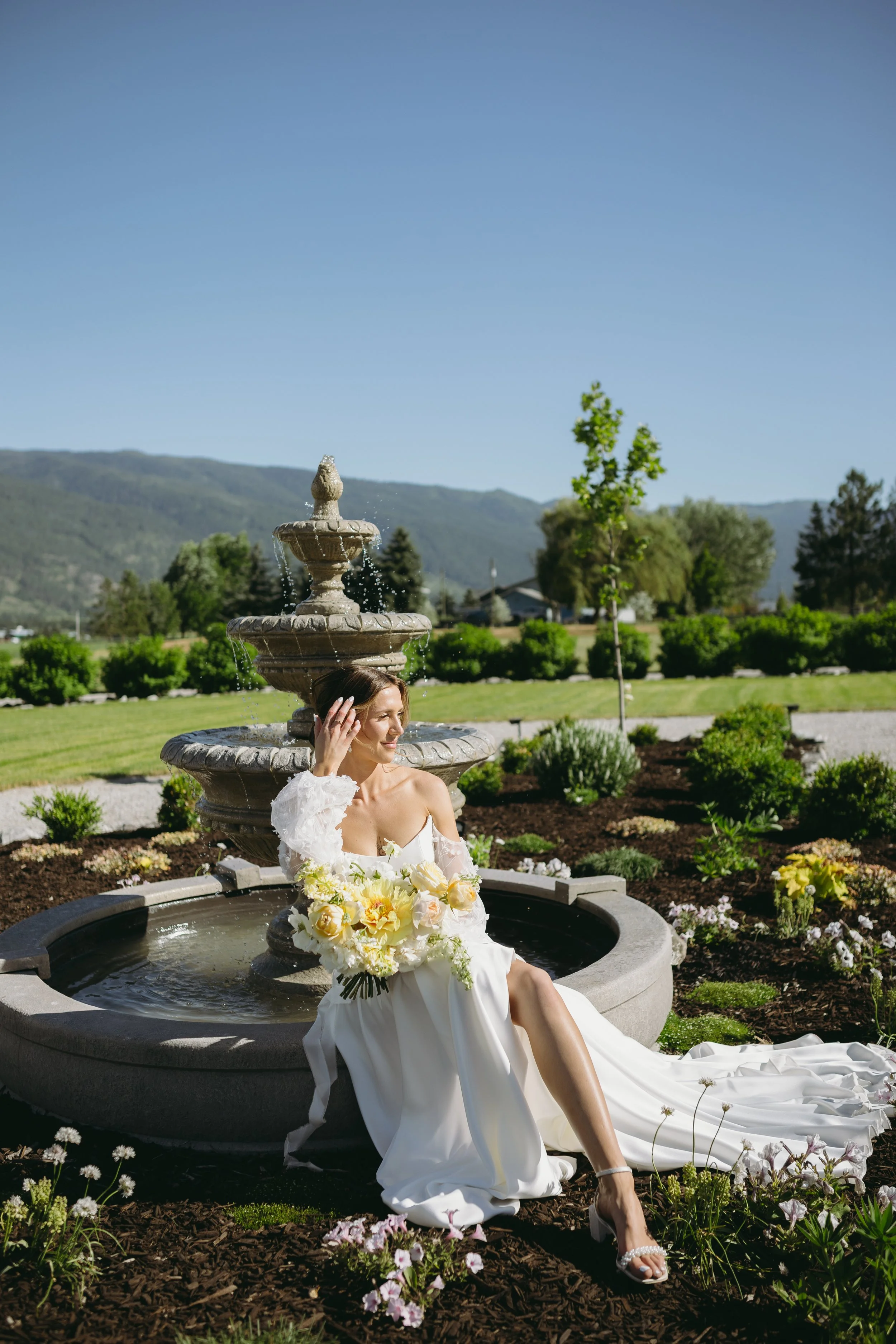 Woman in a white wedding dress sitting by a fountain in a garden.