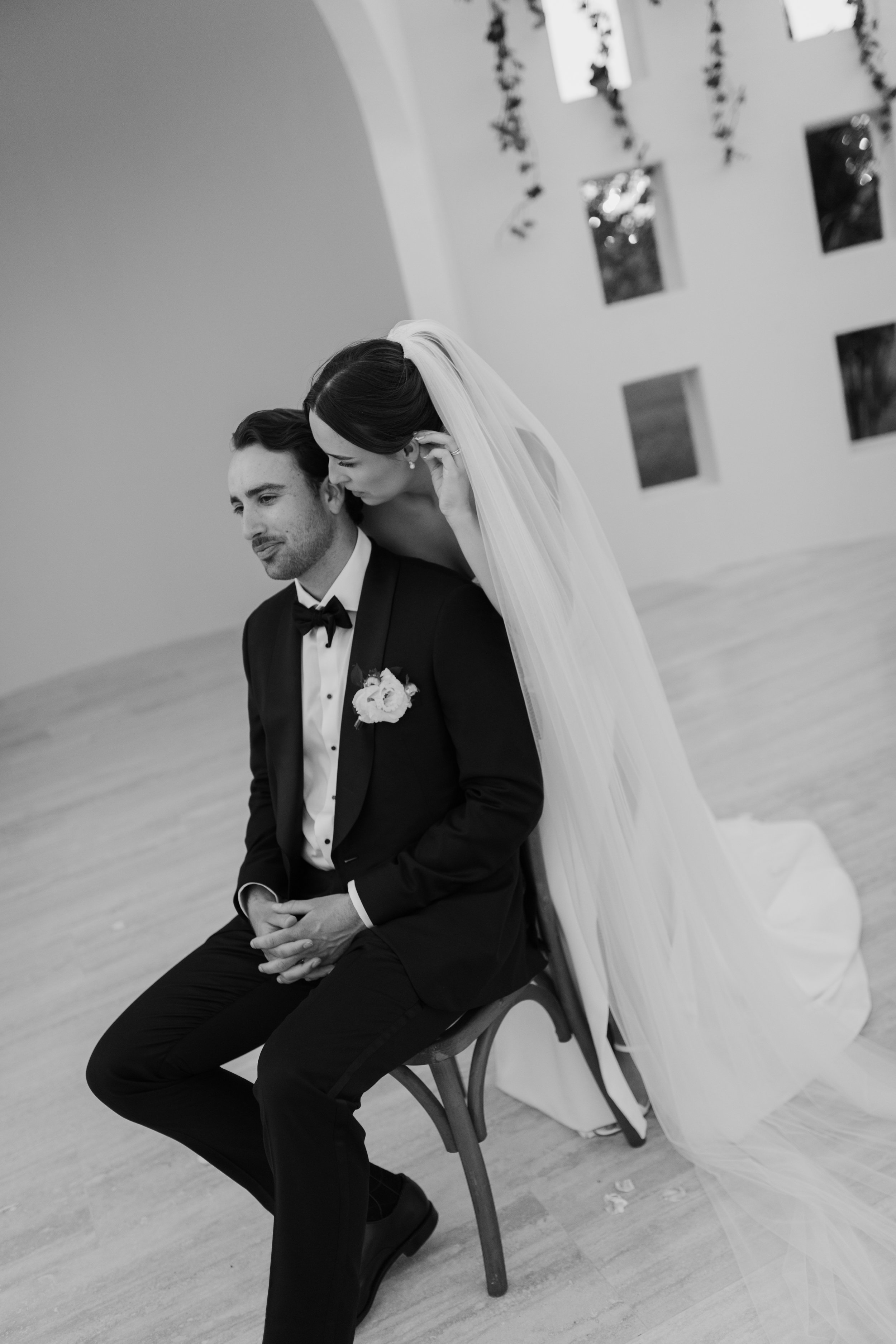Black and white photo of a bride leaning over a seated groom, both dressed in formal wedding attire, in an indoor setting with windows and decorative elements.