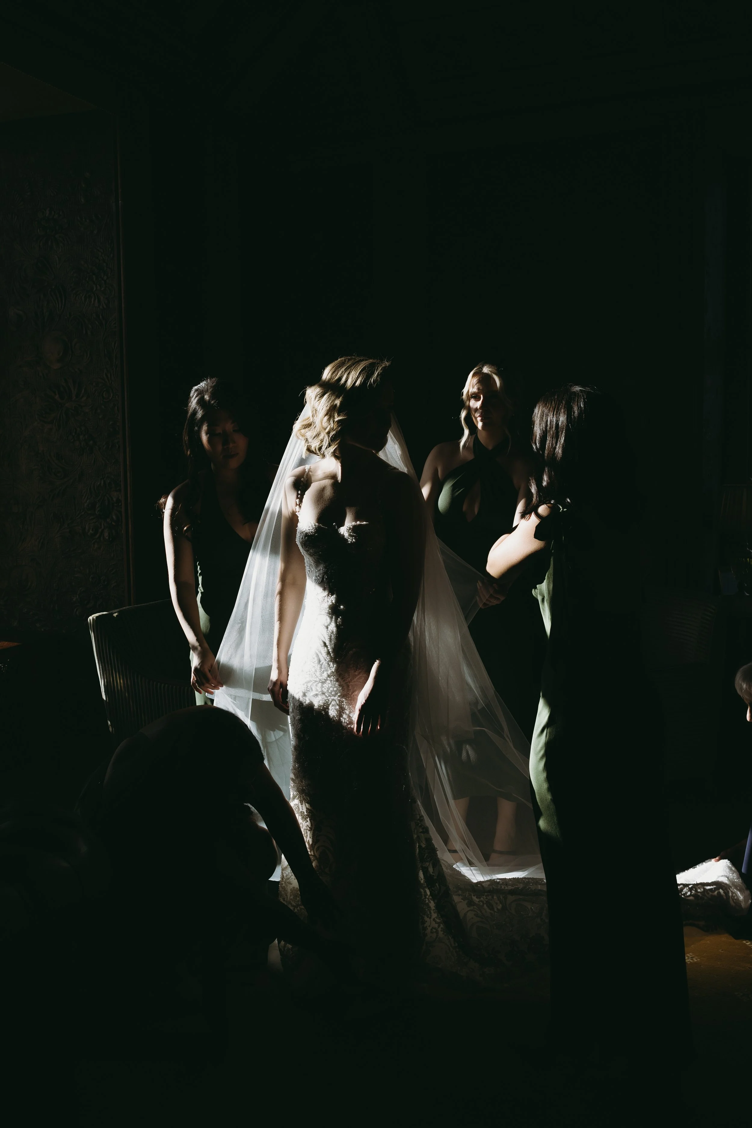 A bride getting ready, silhouetted in dramatic lighting with bridesmaids assisting her.
