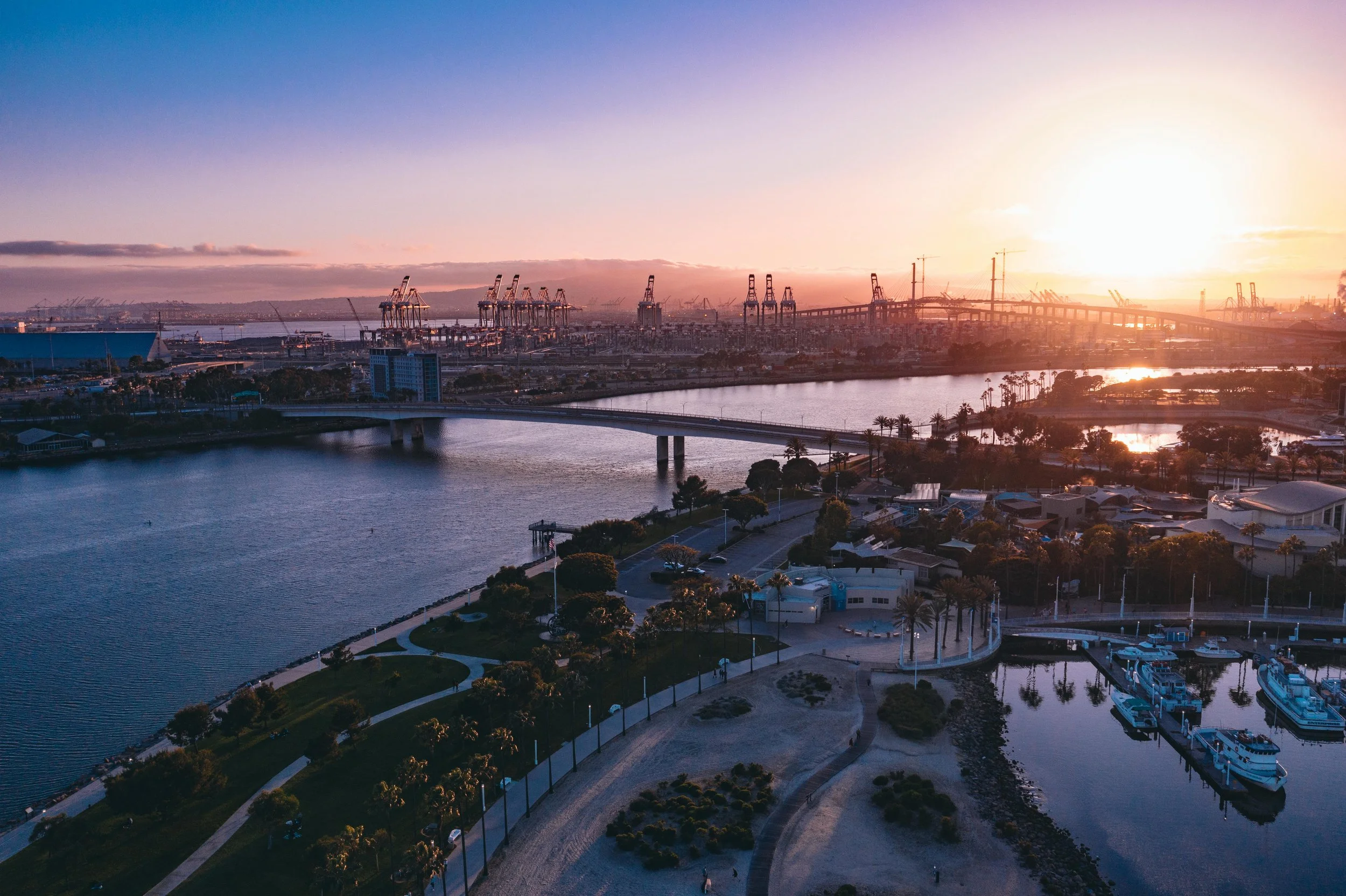 Sunset over a harbor with cranes, bridges, water, and boats, as seen from an elevated viewpoint.