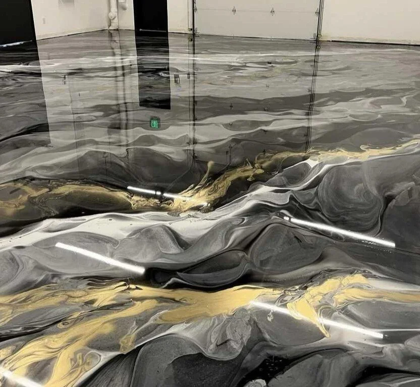 Suelo de piso en un espacio interior, cubierto con una sustancia de apariencia similar a pintura derramada, con tonos negros, grises, dorados y blancos, creando un patrón marbled.