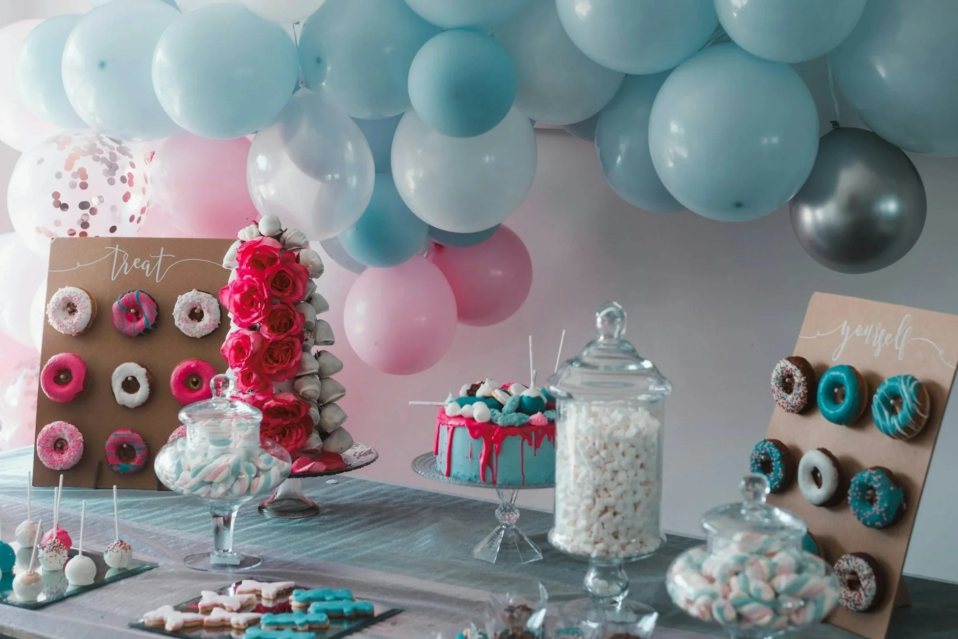 A dessert table decorated with pastel balloons, donuts, cupcakes, and candies for a celebration.
