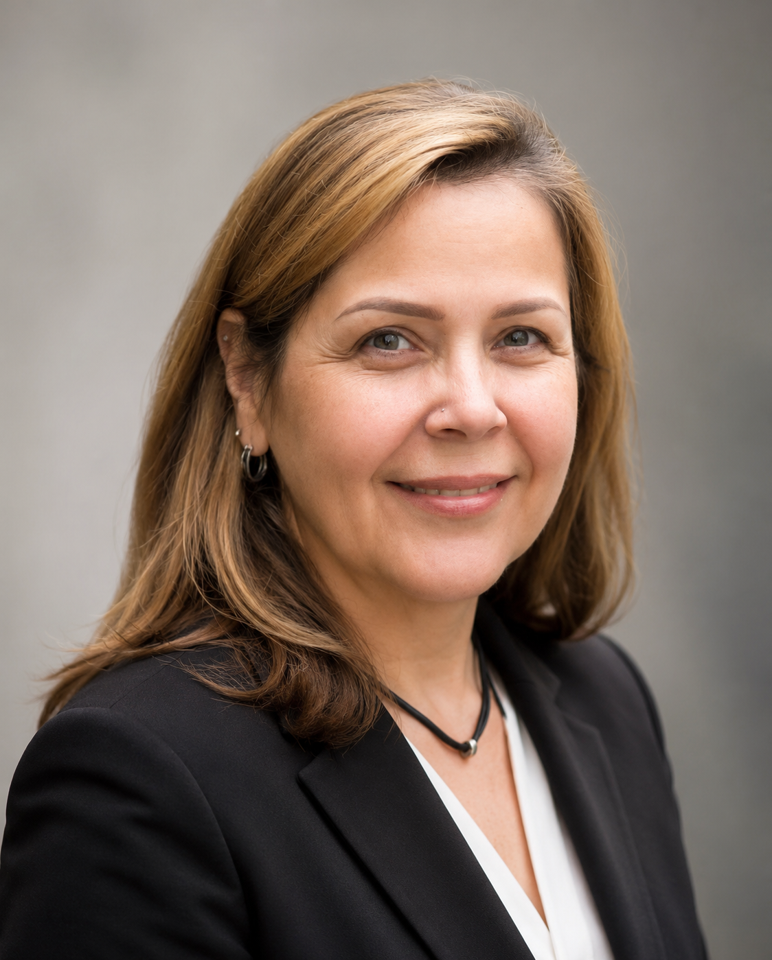 Businesswoman with shoulder-length brown hair wearing a black blazer, white blouse, and jewelry, smiling against a gray background.