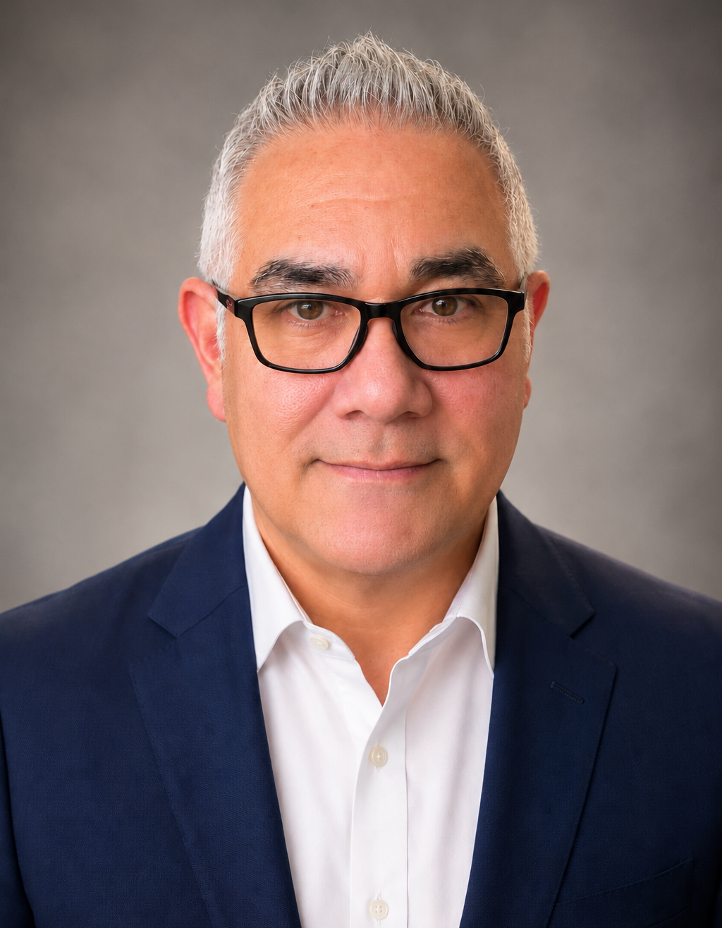Professional headshot of a middle-aged man with gray hair, black glasses, wearing a white shirt and navy suit, standing against a plain gray background.