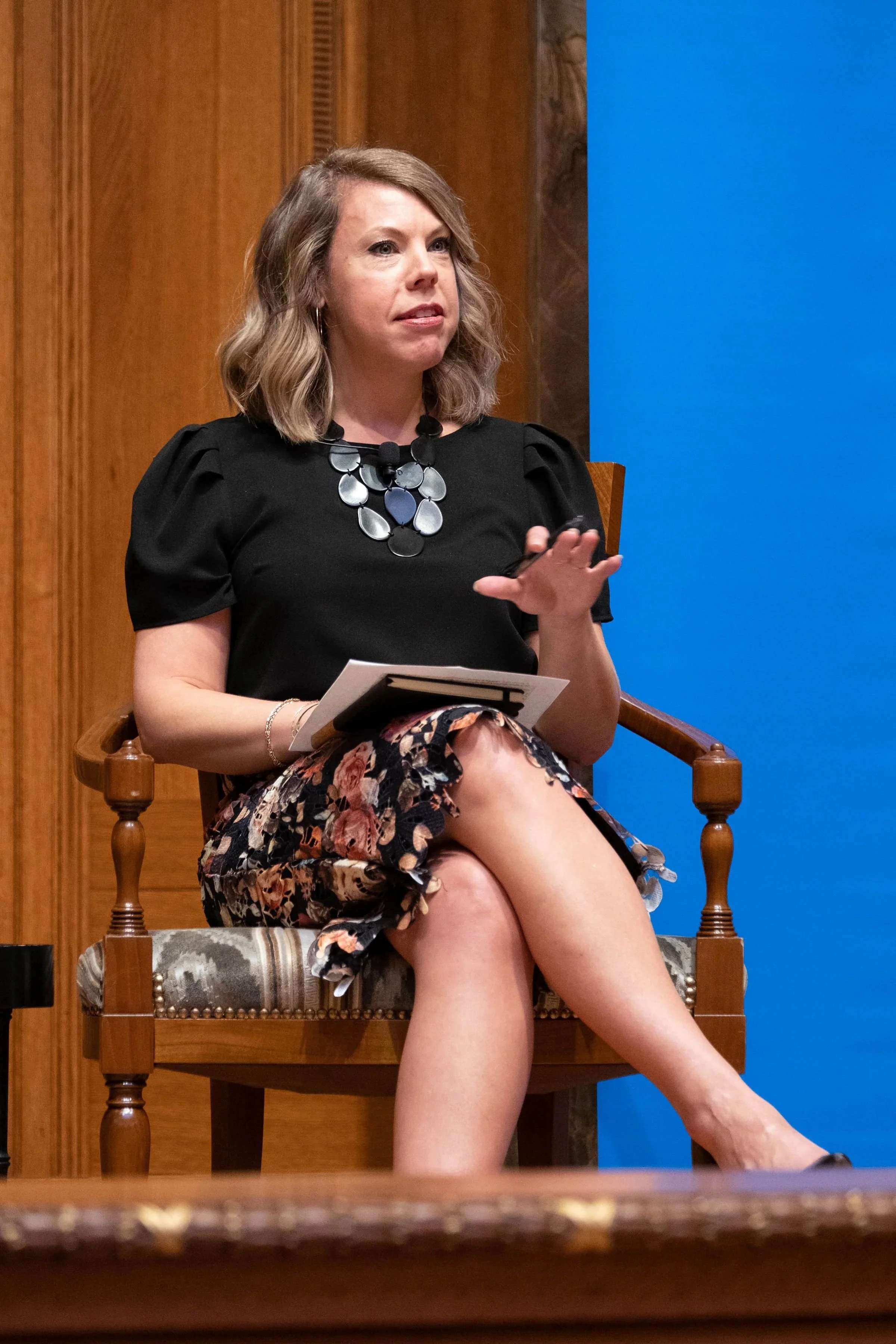 A woman sitting on a wooden chair with a patterned cushion, wearing a black top and floral skirt, holding a notebook, and gesturing with her right hand. Executive coaching for high-performing executives and leaders.