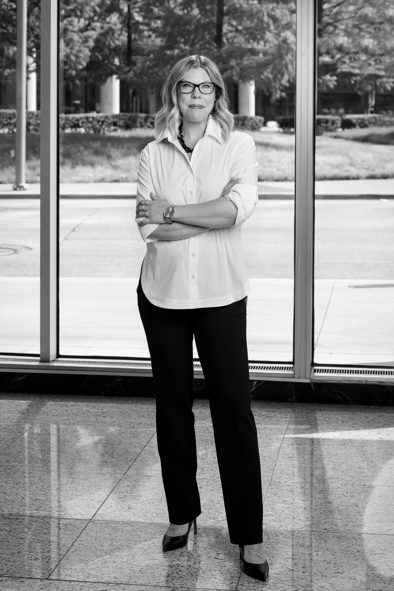 Black and white photo of a woman standing indoors near a large glass window with her arms crossed. She is wearing glasses, a white button-up shirt with rolled-up sleeves, black pants, and high heels. Executive coaching for high-performing leaders.