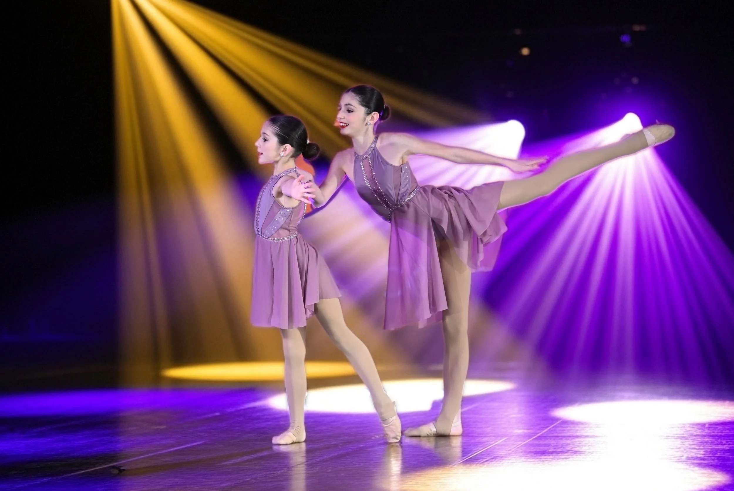 Two young ballet dancers performing on stage under colorful stage lights, one in a pose with arms extended and the other standing with arms crossed.
