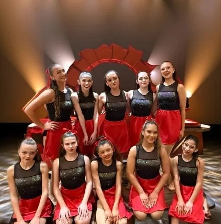 A group of nine young female performers in matching black and red costumes, posing on stage with a red circular backdrop and stage lighting in the background.