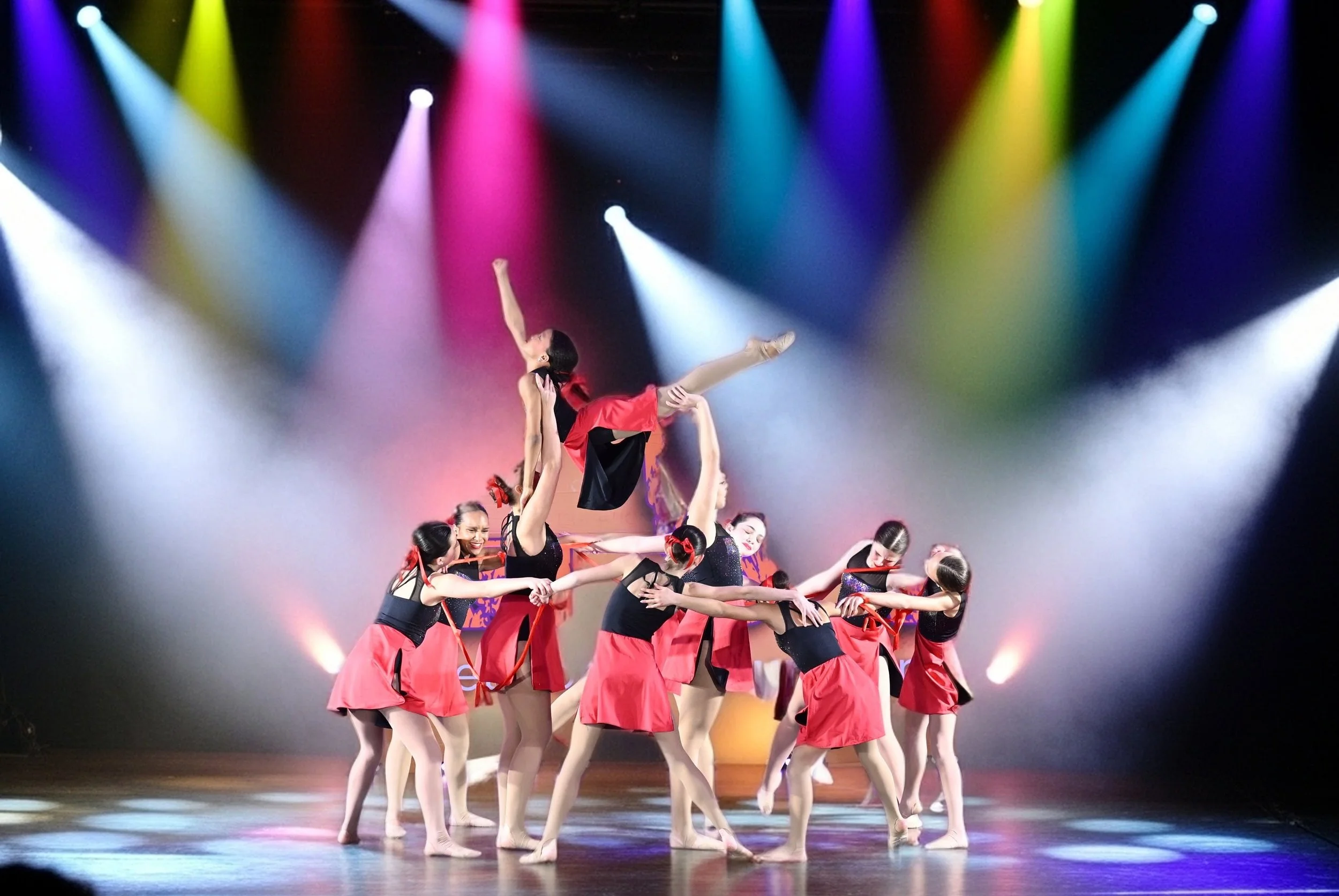 A group of young female dancers performing a ballet routine on stage with colorful spotlights.