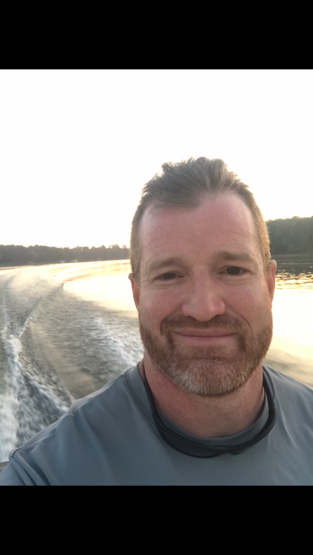 A smiling man taking a selfie on a boat during sunset with water and treeline in the background.
