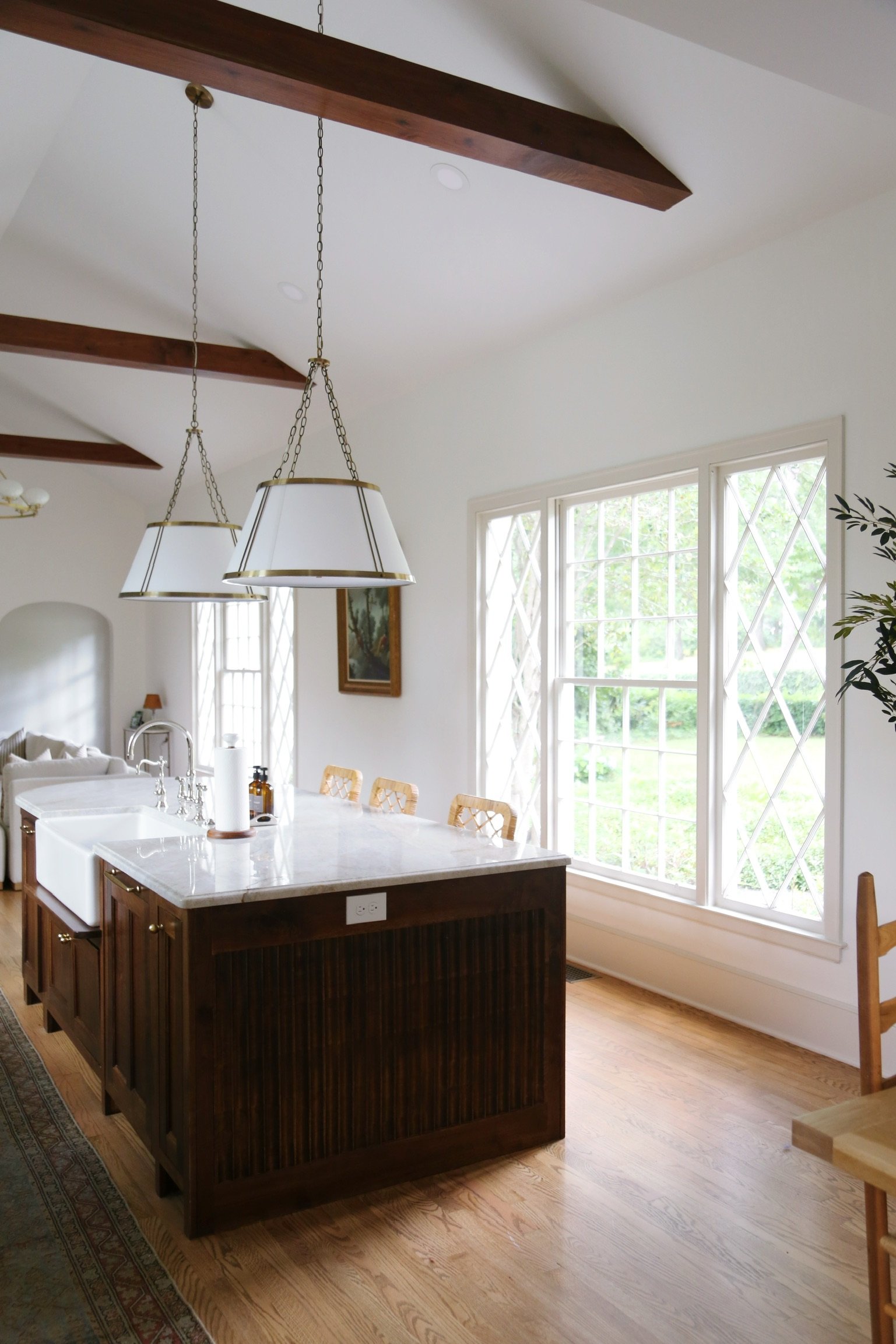 A kitchen with a white island counter, a farmhouse sink, and wooden cabinets, located in a room with large windows and exposed wooden beams on the ceiling, natural light coming through.