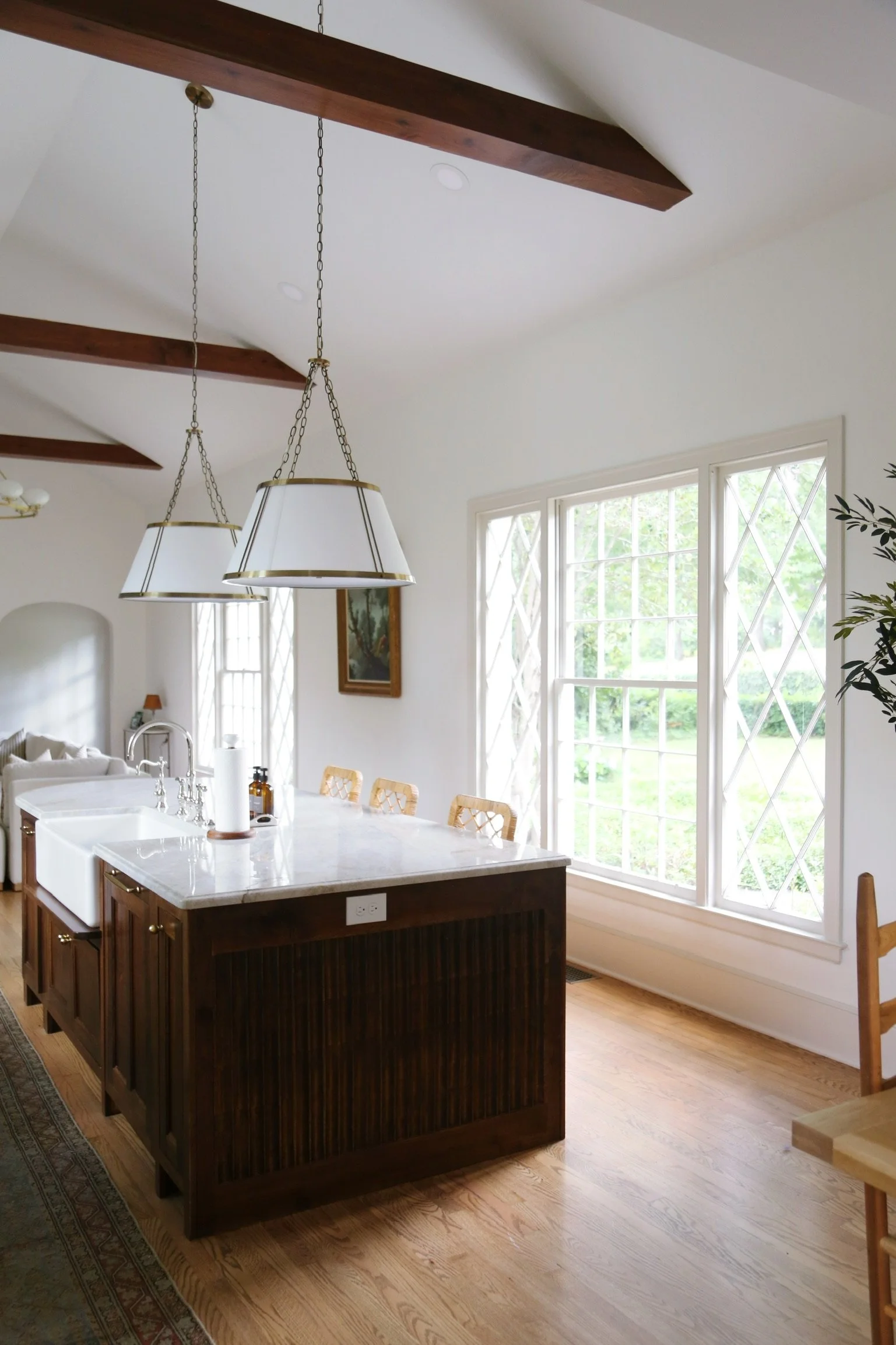 A bright kitchen with a white island counter, dark wood cabinetry, large windows letting in natural light, pendant lights, and hardwood floors.
