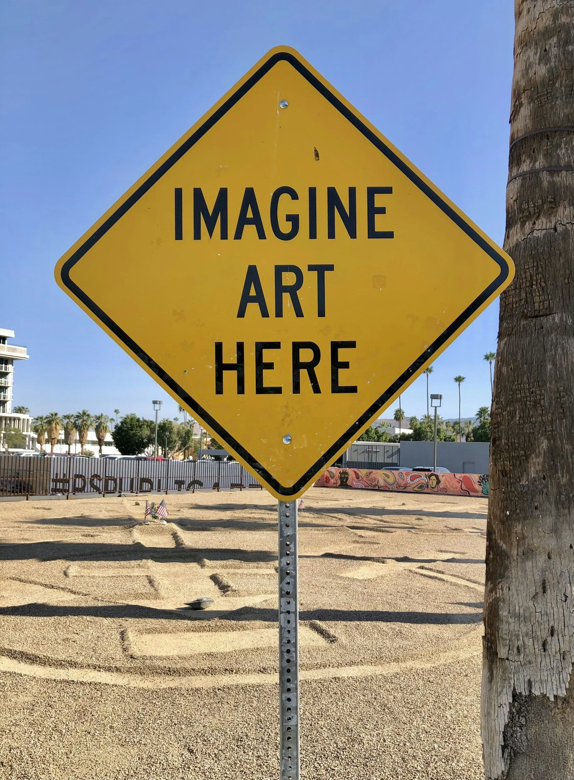 Yellow diamond-shaped sign that reads 'IMAGINE ART HERE' is attached to a metal pole. A tree trunk is on the right. In the background, there are trees, a fence, and buildings.