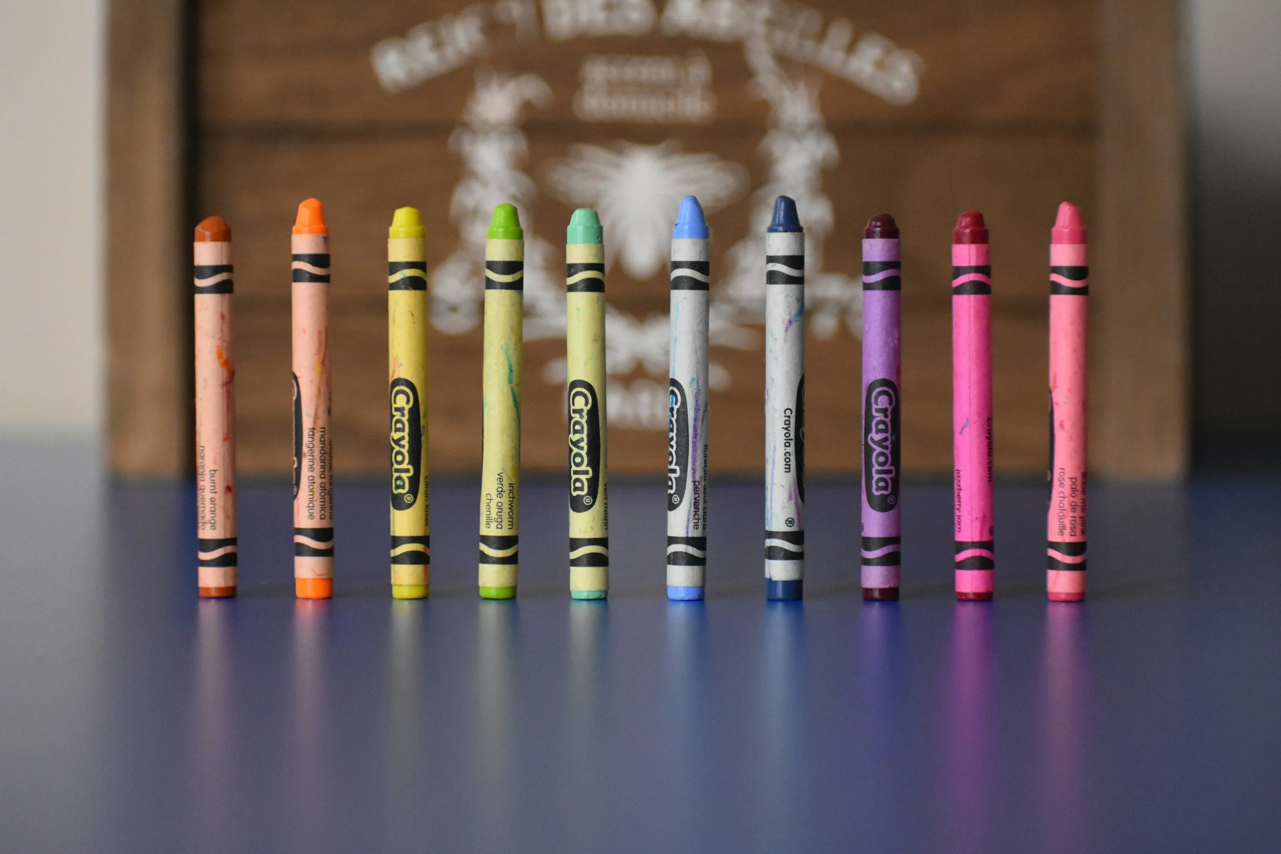 Ten colorful crayons standing upright on a black surface, with a blurred background featuring a decorative skull design.