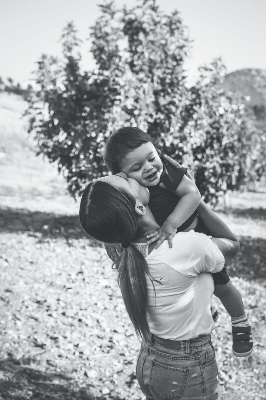 A woman lifting a smiling young boy outdoors in front of a tree.