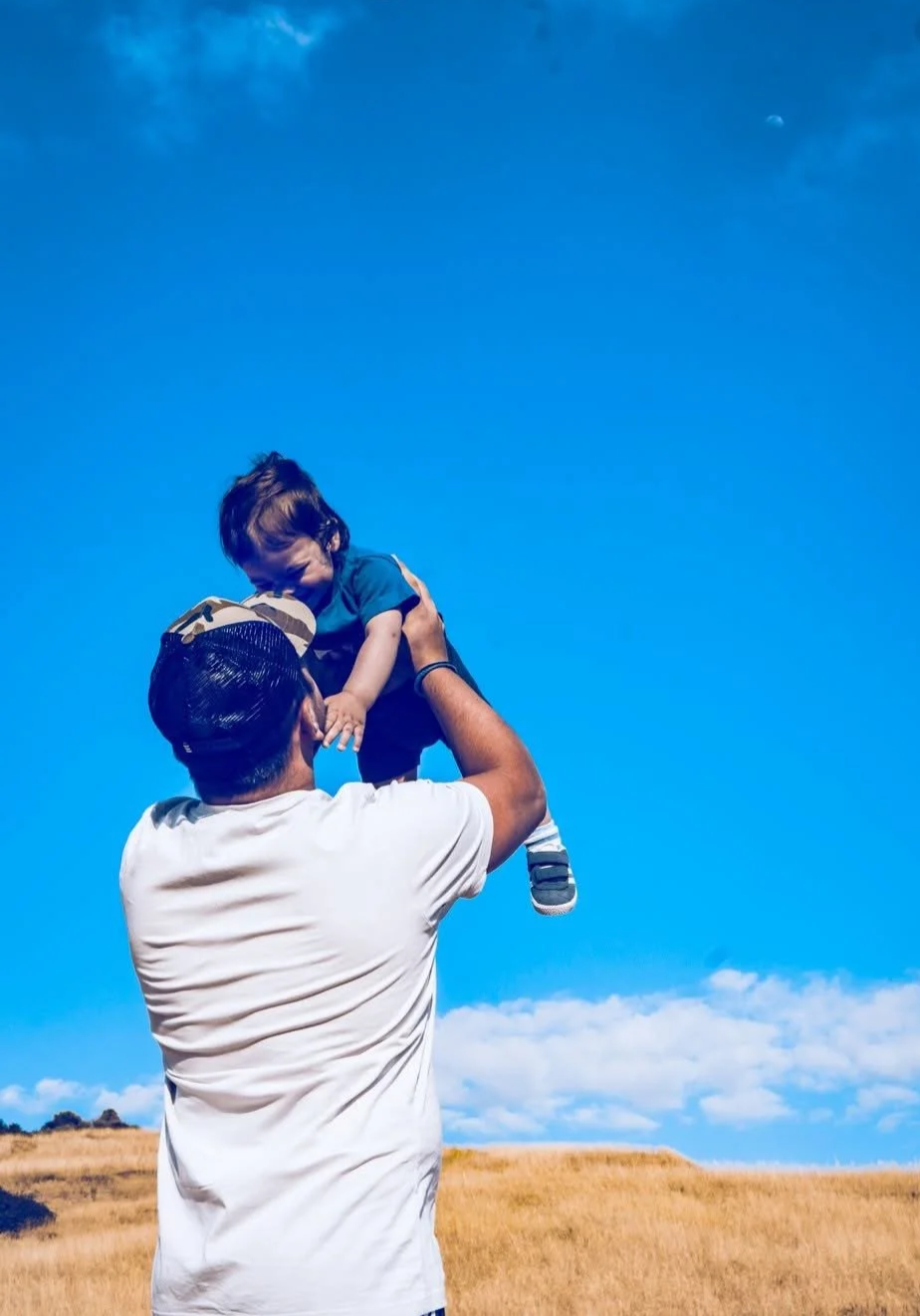 A man lifting a young boy into the air outdoors under a blue sky with some clouds and the moon visible.