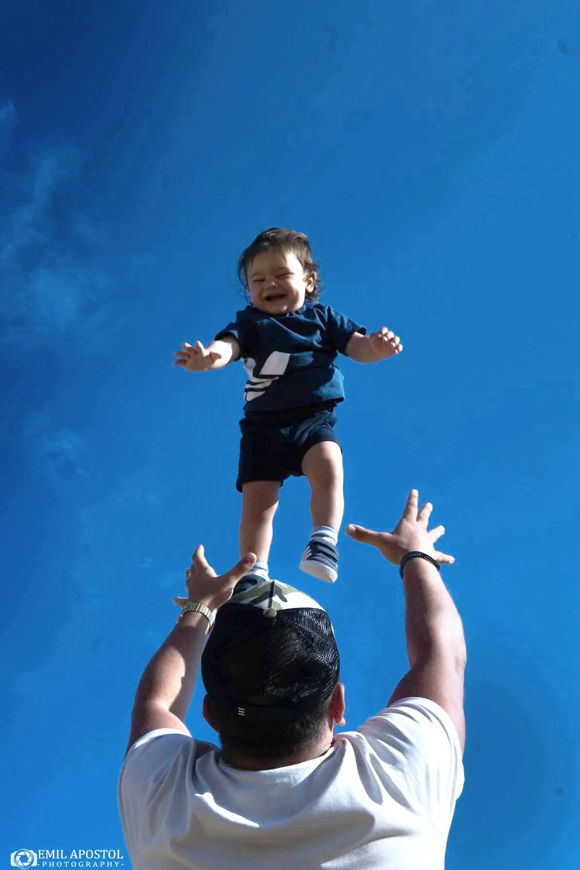 A man in a white shirt and black cap lifting a young boy in a blue outfit into the air against a blue sky.