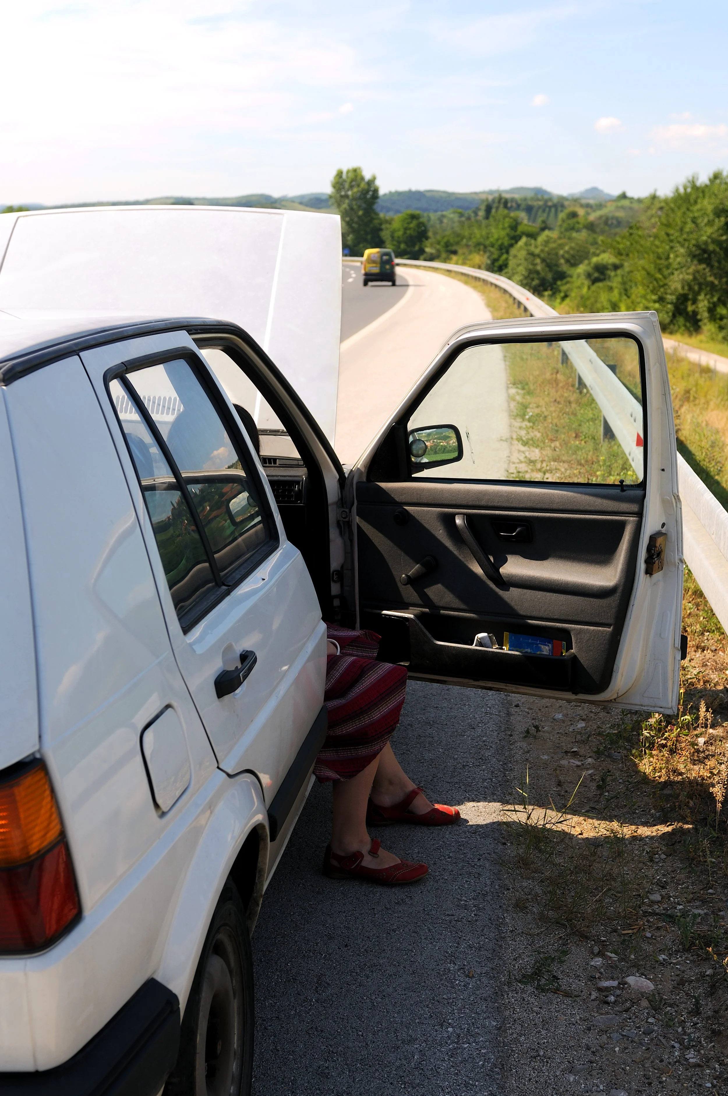 A person sitting in a white car on the side of a rural road, with the car door open and the hood raised, surrounded by green trees and rolling hills under a blue sky with some clouds.